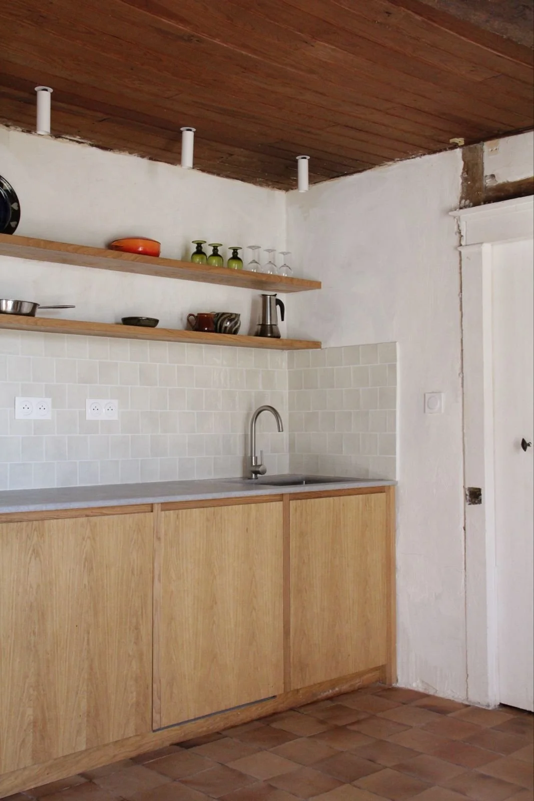 A minimalist kitchen with wooden cabinets, a single sink, open wooden shelves with dishes and glasses, and a wooden ceiling.