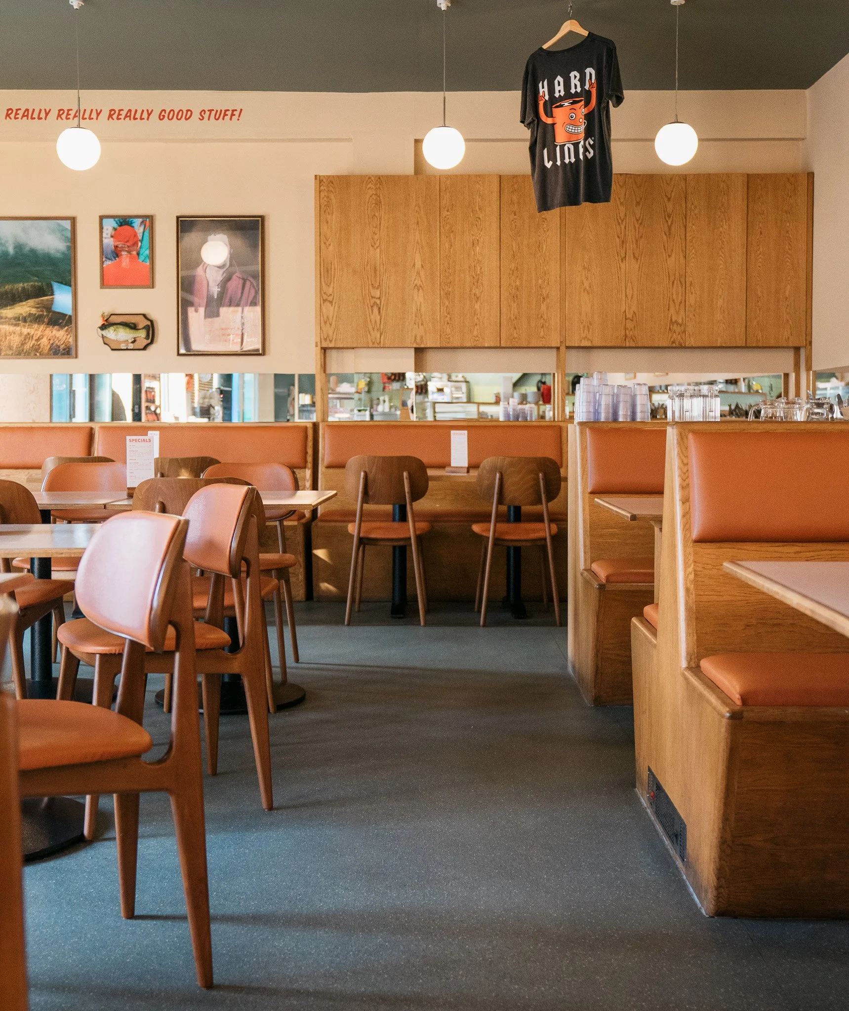 Empty dining area with wooden chairs and booths, ceiling lights, and wall art, including a shirt hanging with a graphic and the words 'HARD LIONS'.
