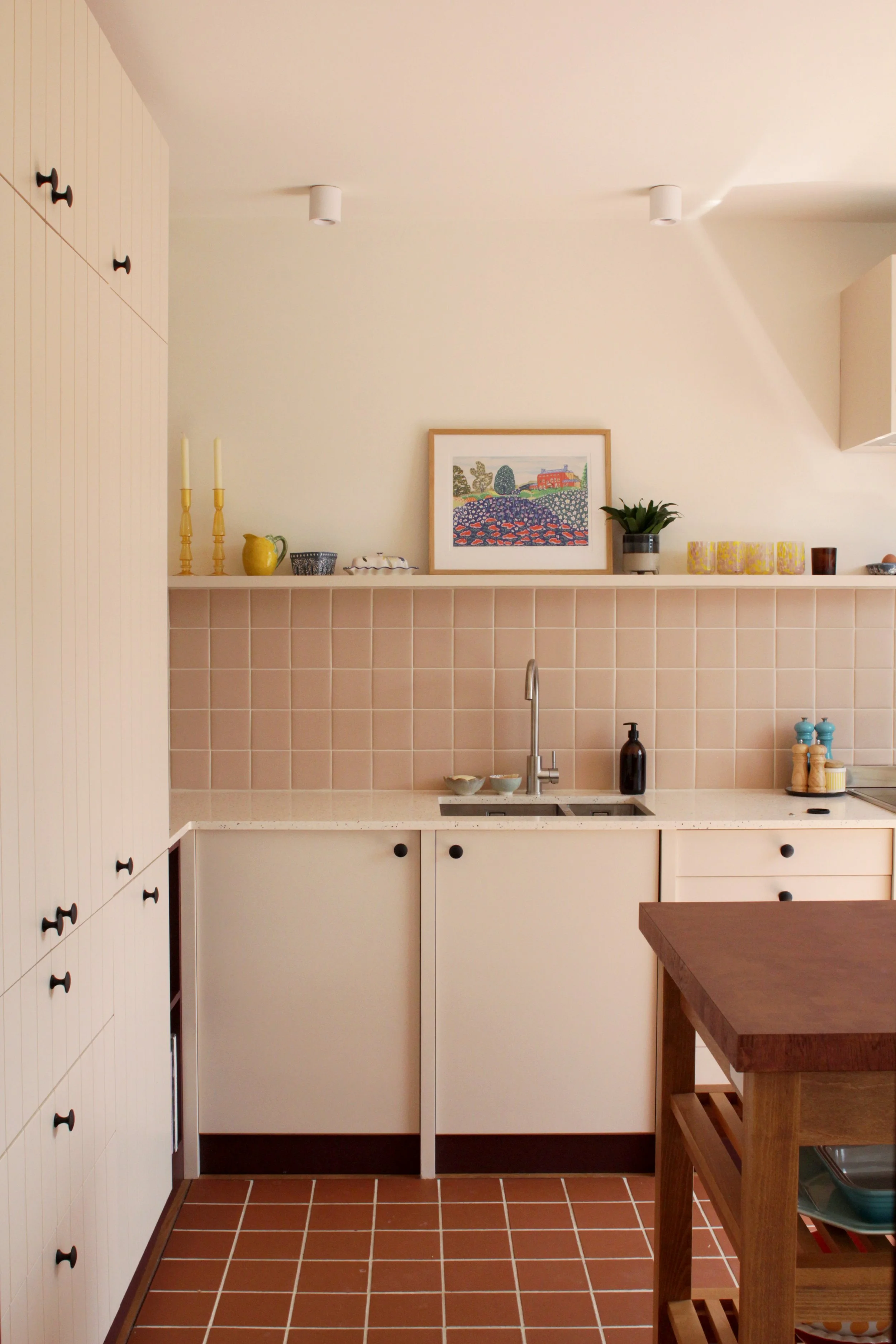 A minimalist kitchen with white cabinets, a white countertop, a beige tiled wall, and a wooden kitchen island. Decor items include a framed picture, plants, candles, and spice jars.