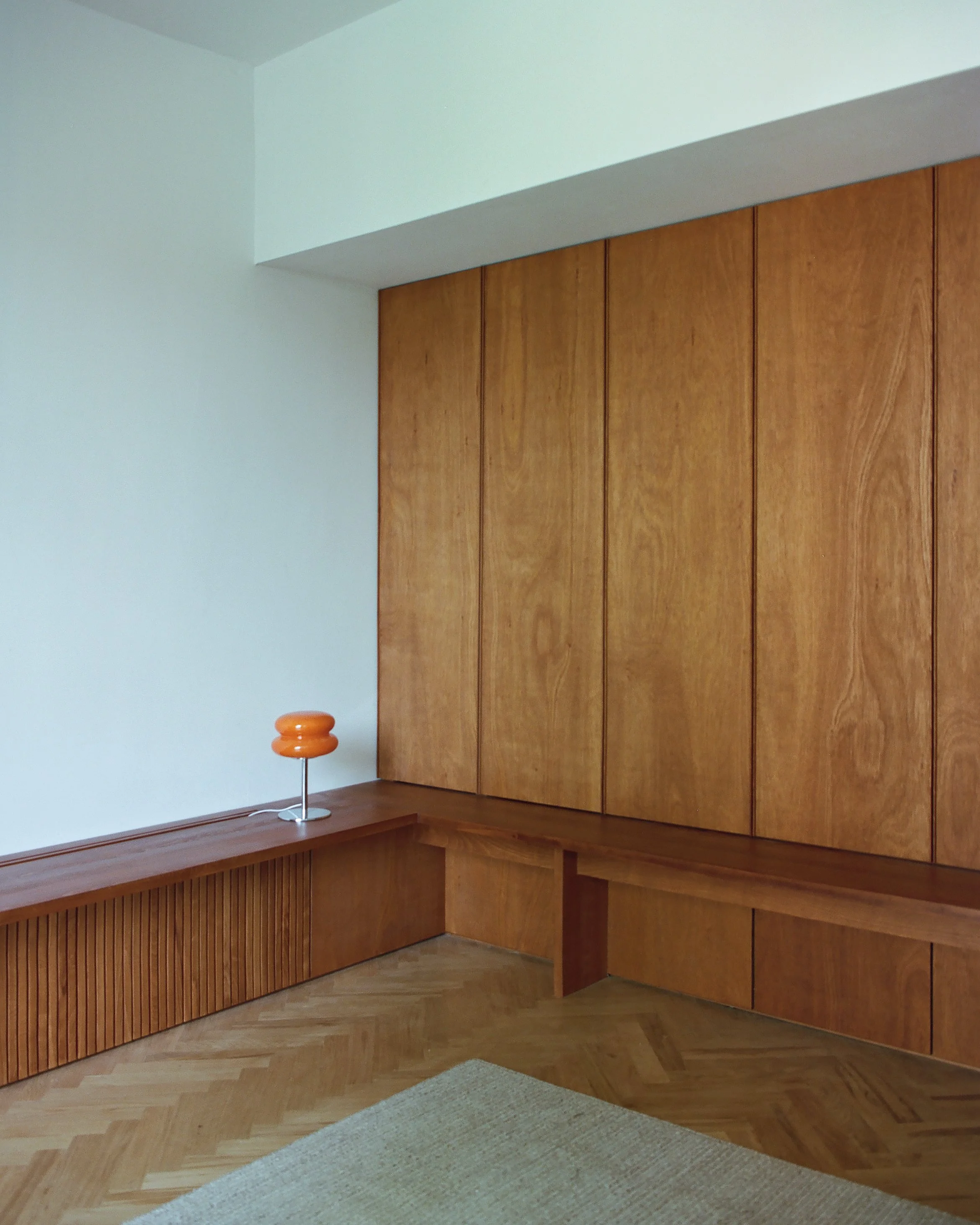 Corner of a room with wooden built-in cabinets and a wooden bench, with a white wall and a small orange lamp on the bench.