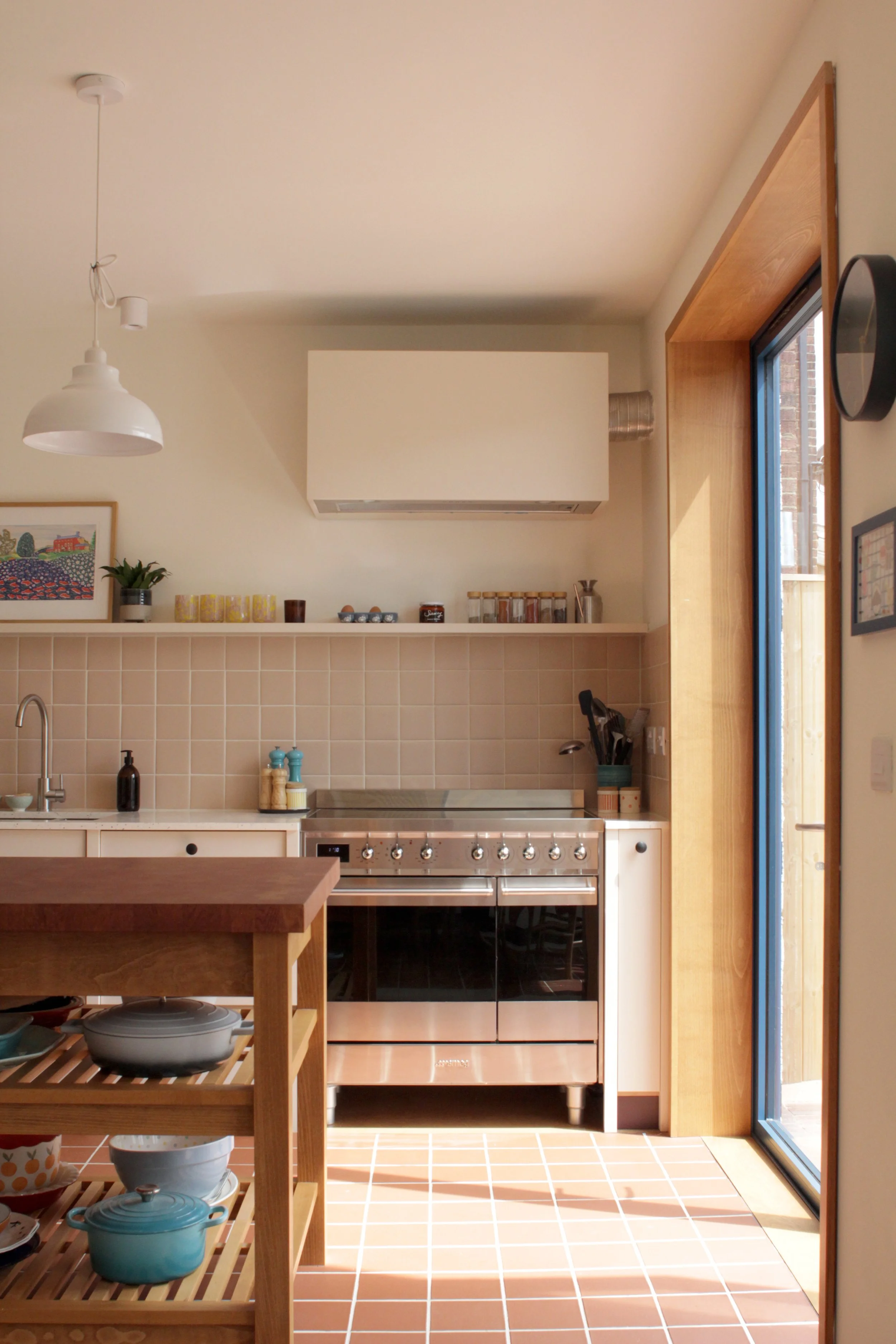 Bright, minimalist kitchen with beige tile backsplash, stainless steel stove, white cabinetry, and wooden accents. Sunlight streams in through a glass door.