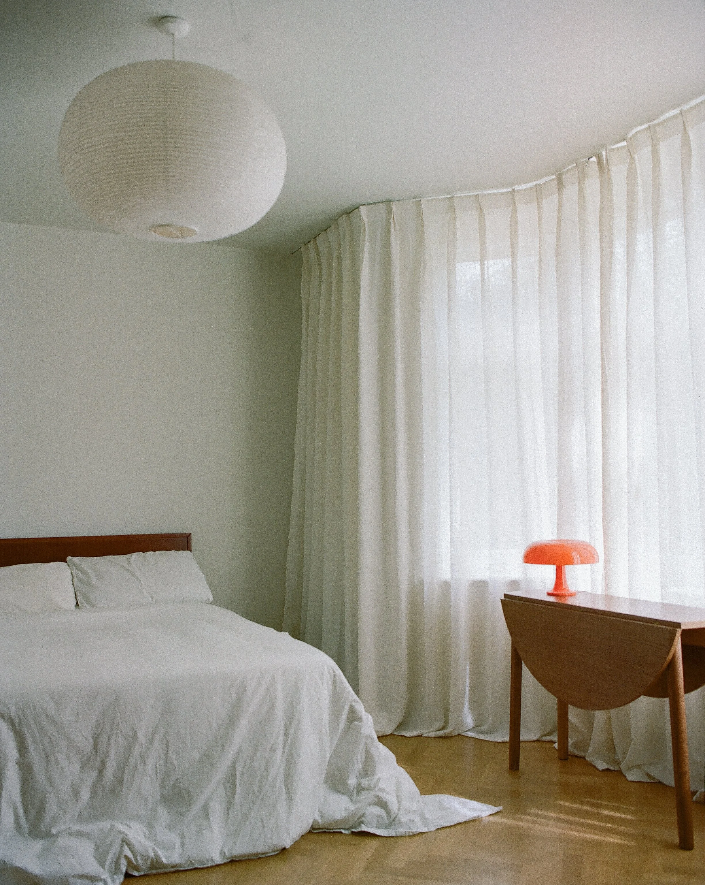 A bedroom with a bed covered in white linens, a wooden nightstand with an orange table lamp, cream curtains, a wooden floor, and a white paper lantern ceiling light.