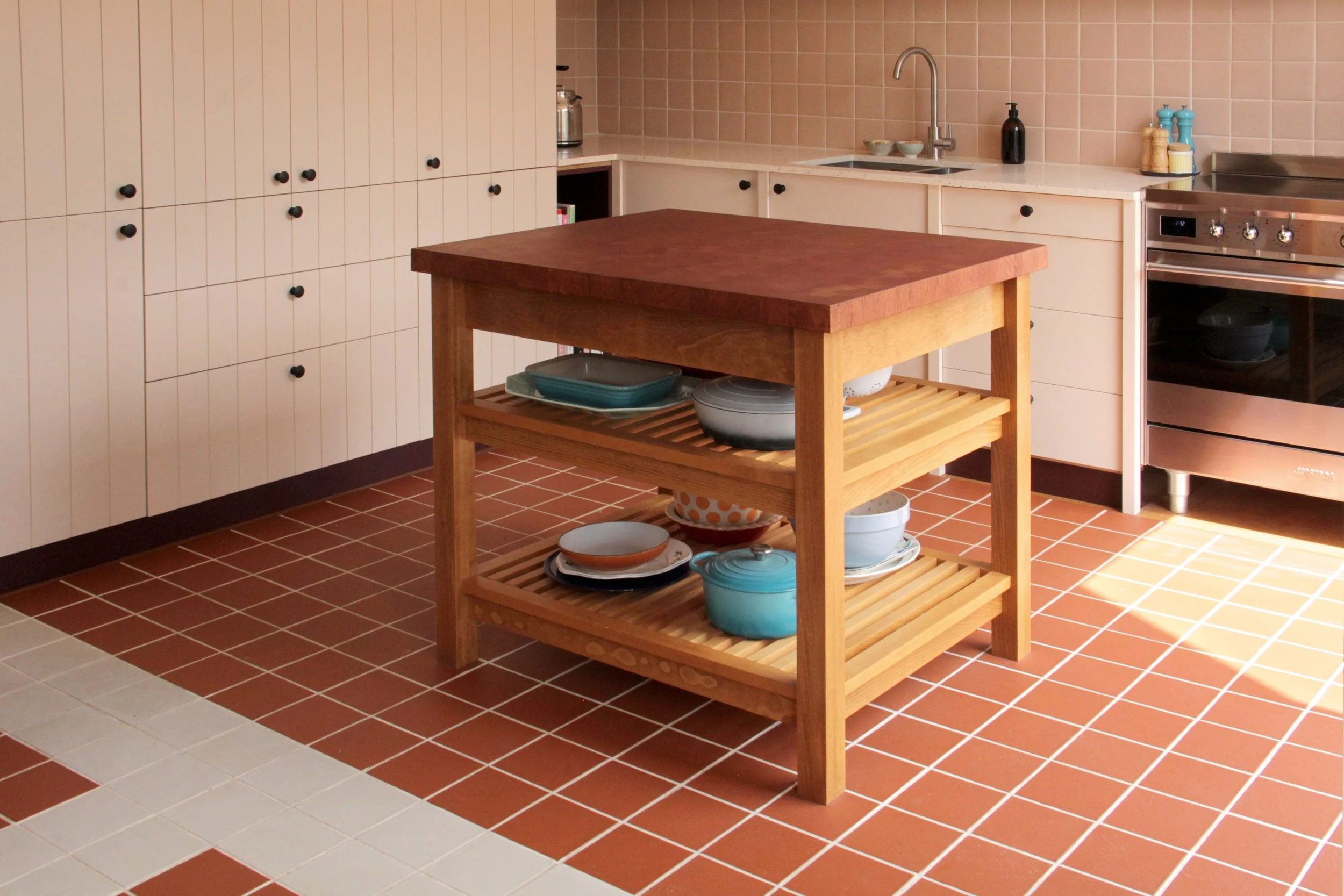 A kitchen with white cabinets, a brown tiled floor, and a central wooden kitchen island with open shelving holding plates and bowls.