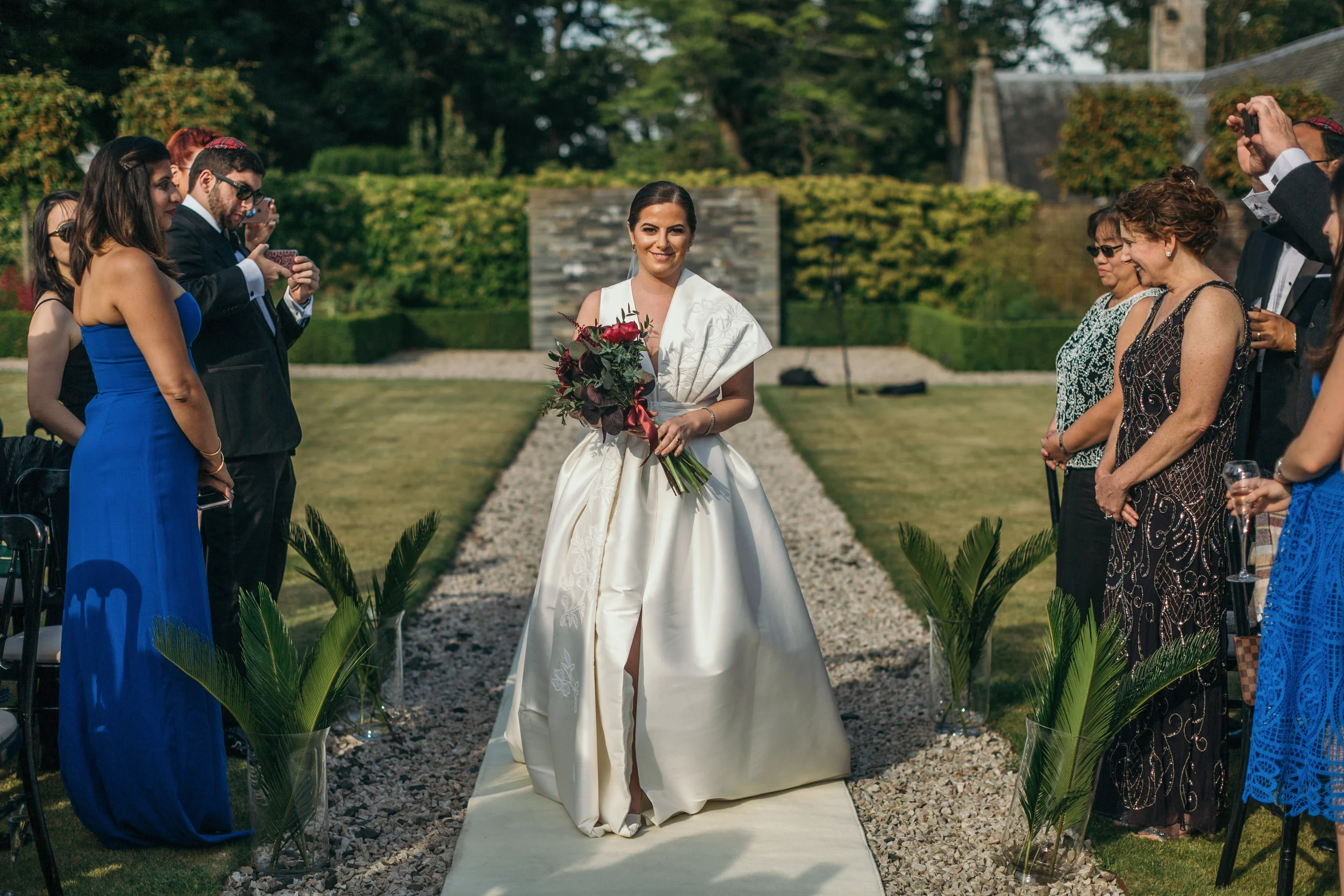 Beautiful bride walking down aisle with organic wine flower bouquet wearing ivory one shoulder wedding dress in outdoor castle ceremony