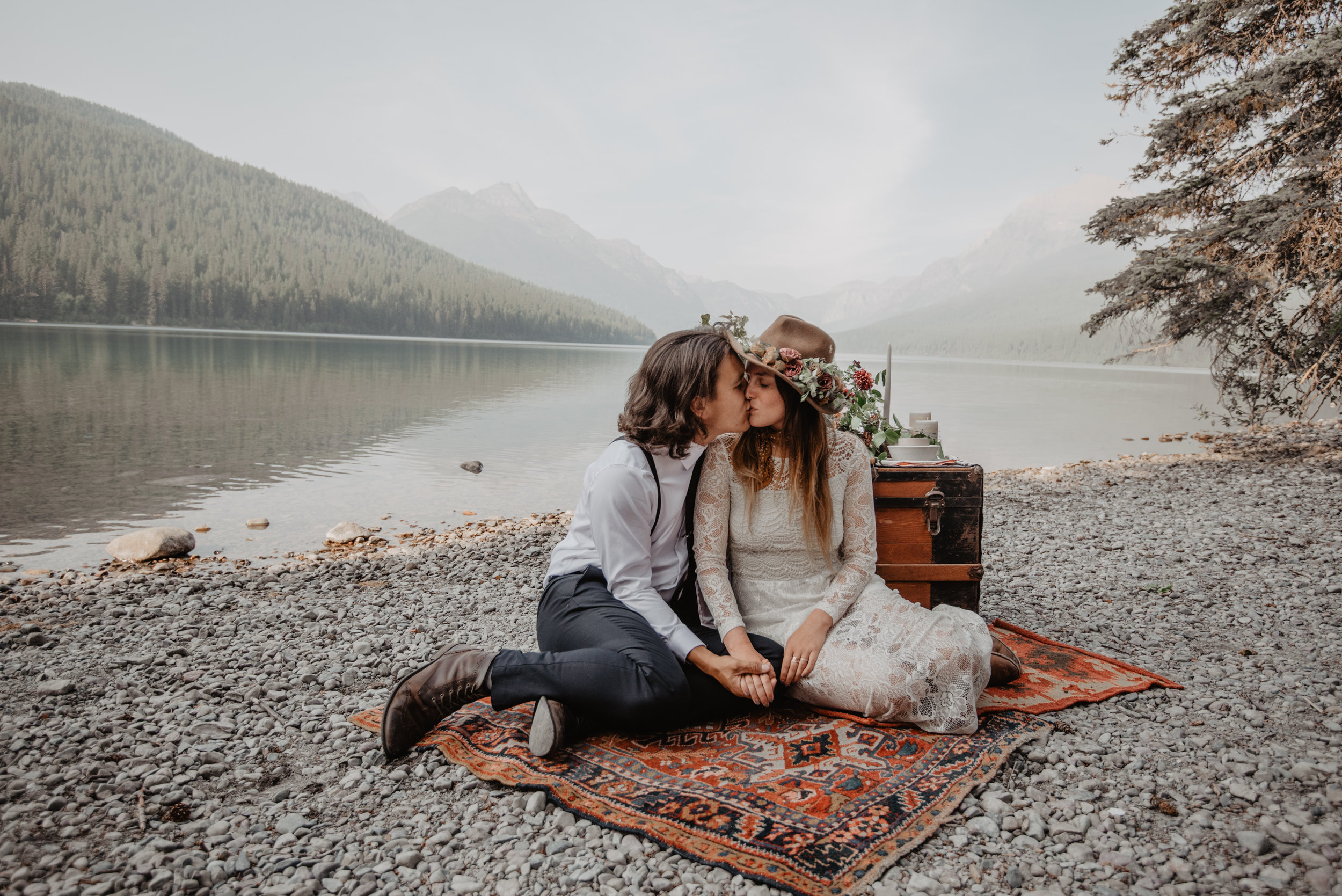 Outdoor elopement portrait of engaged couple sitting on renewable and sustainable bohemian carpet while kissing each other at Glacier National Park MT
