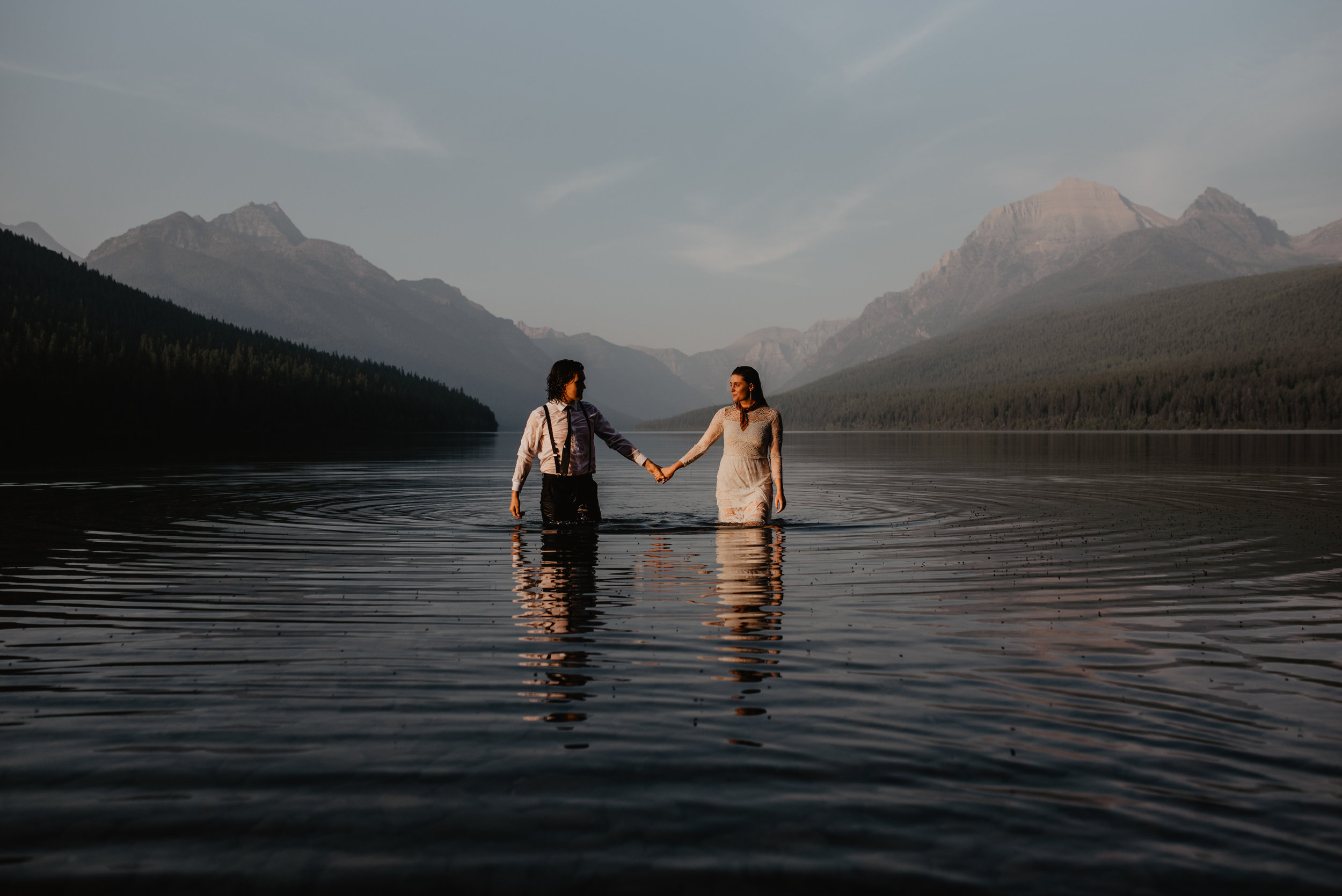 Outdoor elopement portrait of happy engaged couple holding hands in water at Glacier National Park MT