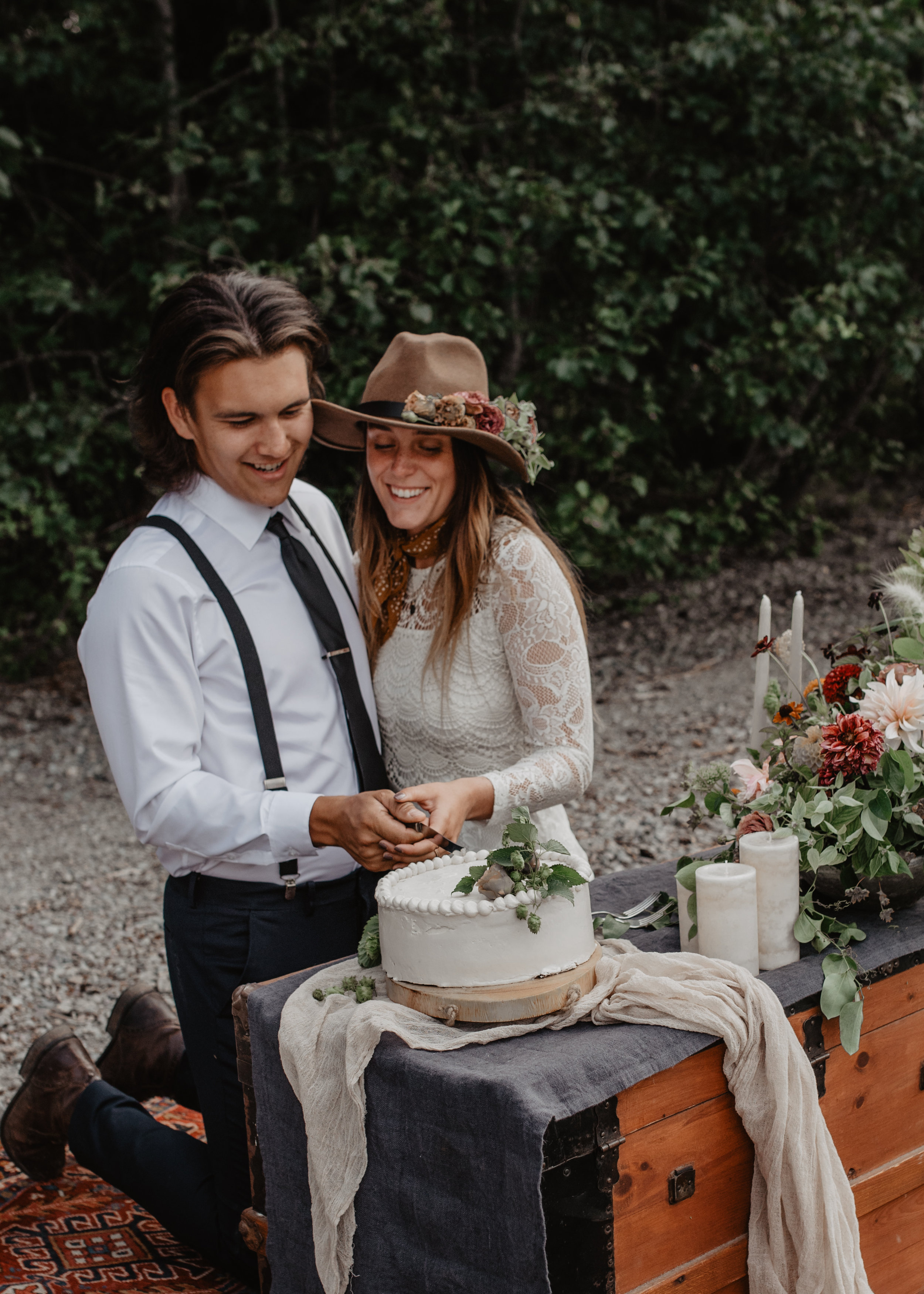 Outdoor elopement portrait of engaged couple kneeling on renewable and sustainable bohemian carpet cutting their local cake at Glacier National Park MT