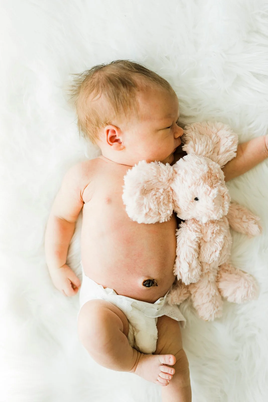 A sleeping baby lying on a soft white surface, cuddling a light pink teddy bear.