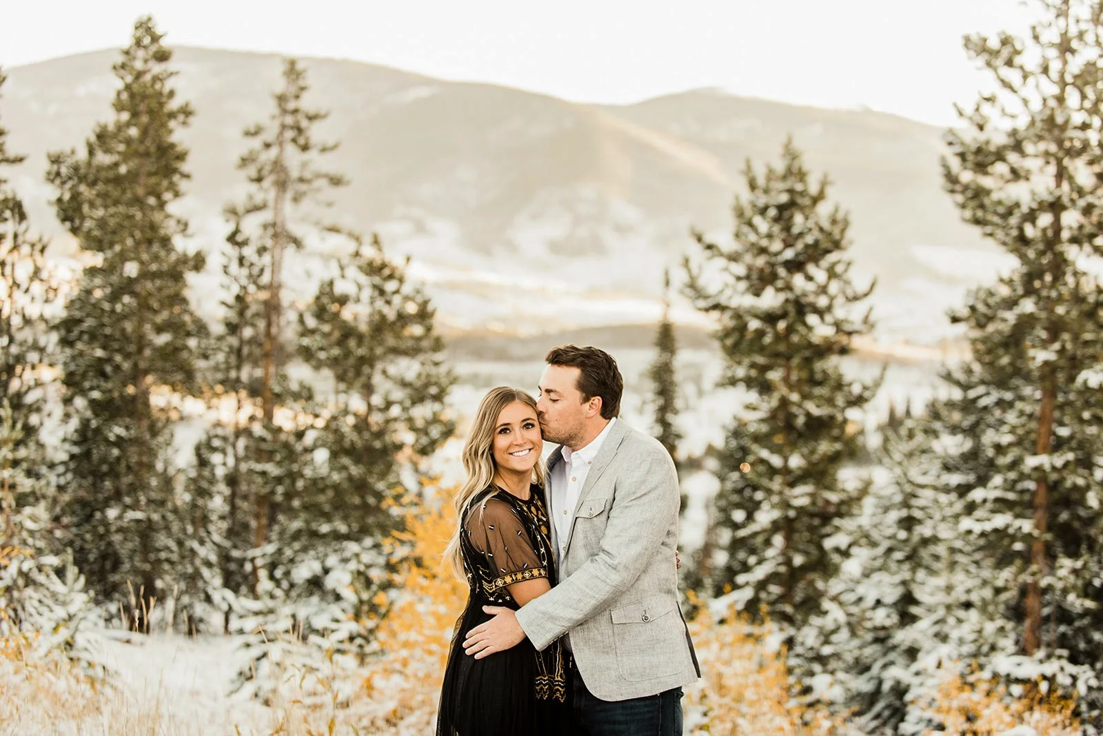 A smiling woman in a black dress with patterned detailing, and a man in a light gray blazer and white shirt, embrace in a snowy forest with evergreen trees and mountains in the background.