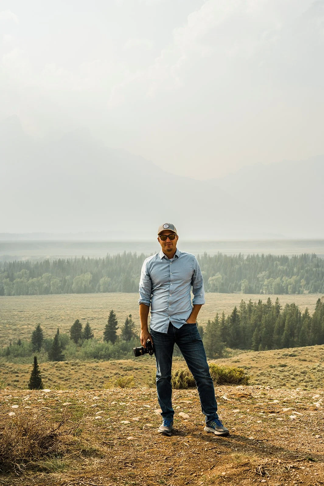 A man standing outdoors on a rocky terrain with a camera in his hand, wearing sunglasses, a light blue shirt, and a baseball cap, with a background of forests and mountains under a cloudy sky.