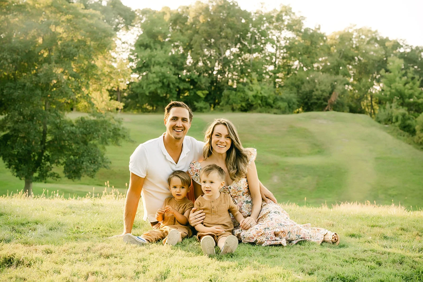 A smiling family of four sitting on grass in a park with green trees in the background during daylight.