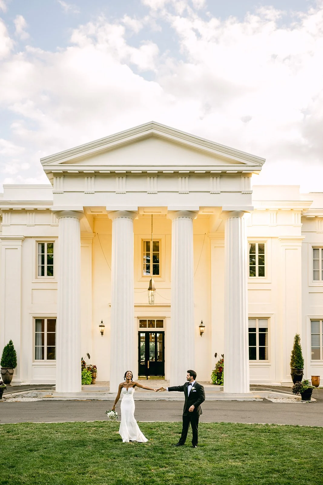 A bride and groom stand on a grassy lawn in front of a large white building with tall columns, holding hands and smiling. The bride wears a white wedding dress and holds a bouquet, while the groom is in a black tuxedo.