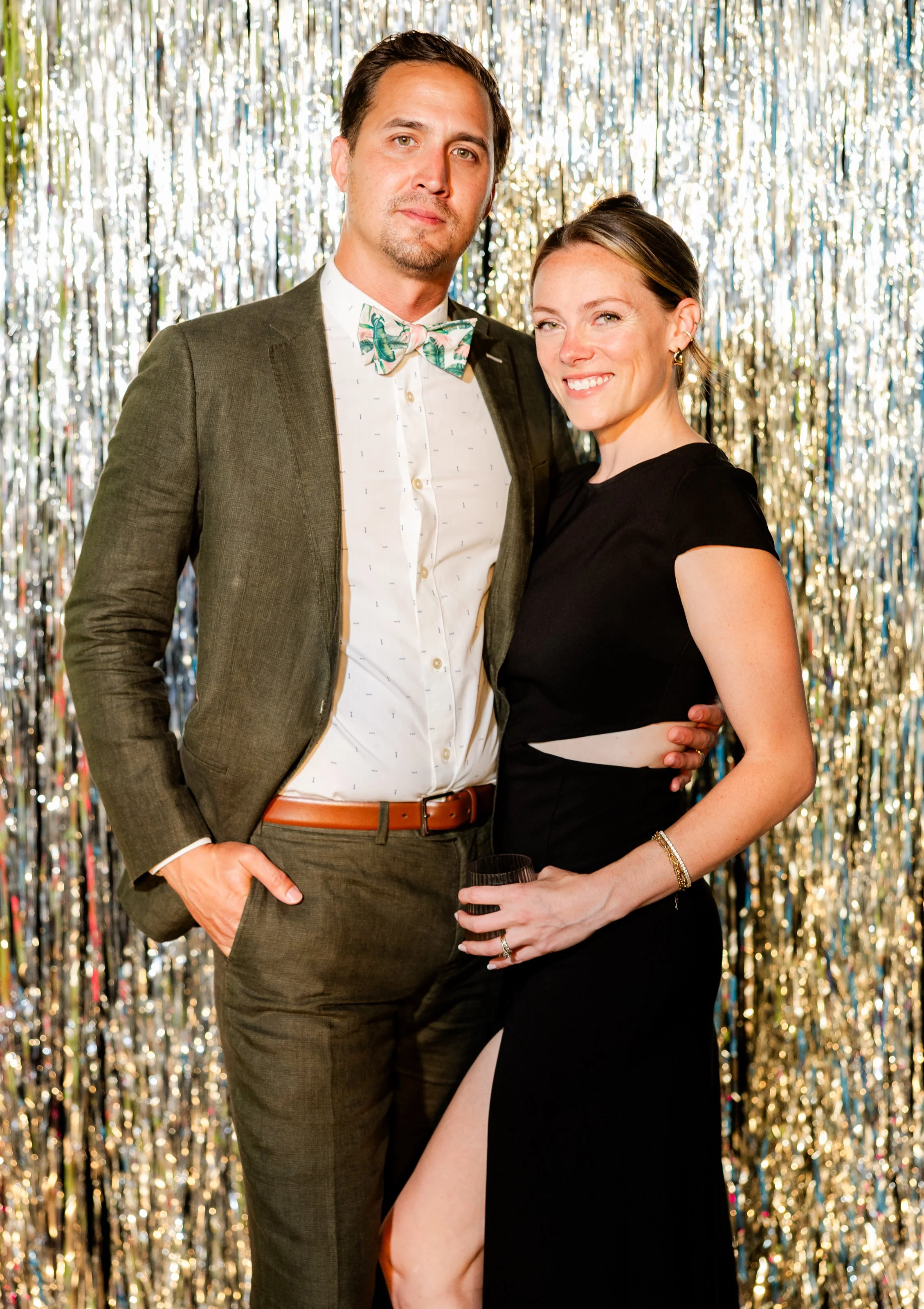 A well-dressed couple posing in front of metallic gold and silver streamers at a party. The man wears a dark green suit, white shirt with a pattern, and a colorful bowtie. The woman wears a black dress with a high slit, holding a glass, and smiling.