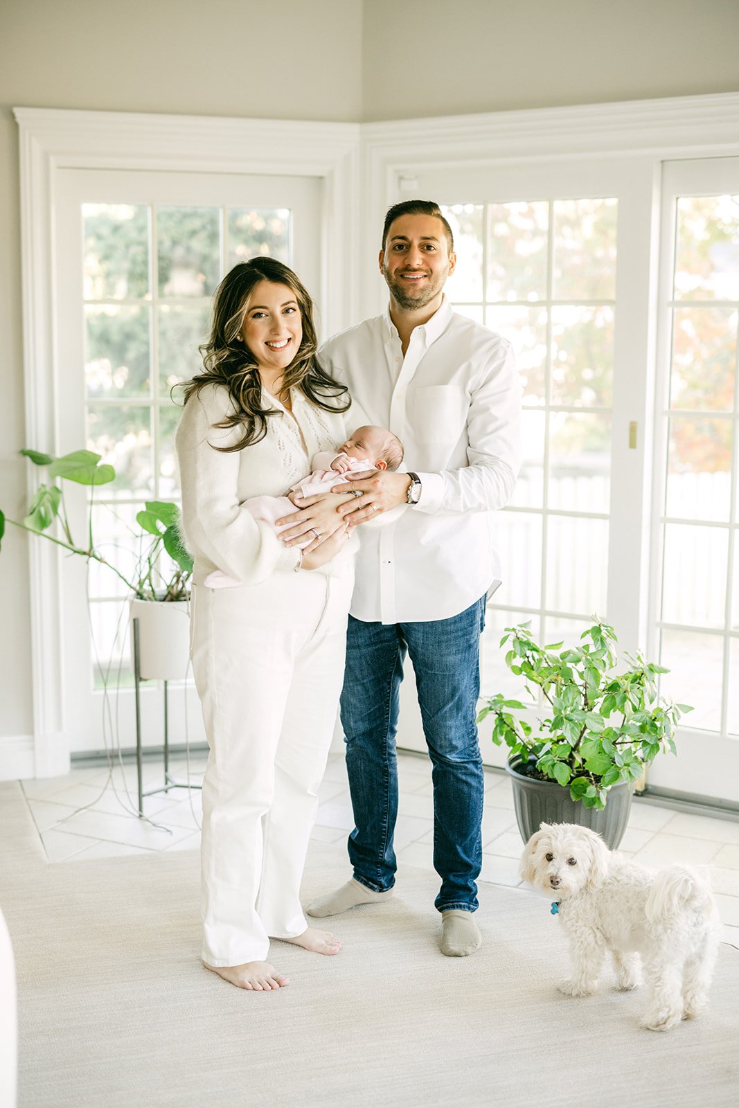 A family of three with their small white dog, standing in a bright room with large windows and indoor plants. The mother and father hold a newborn baby.