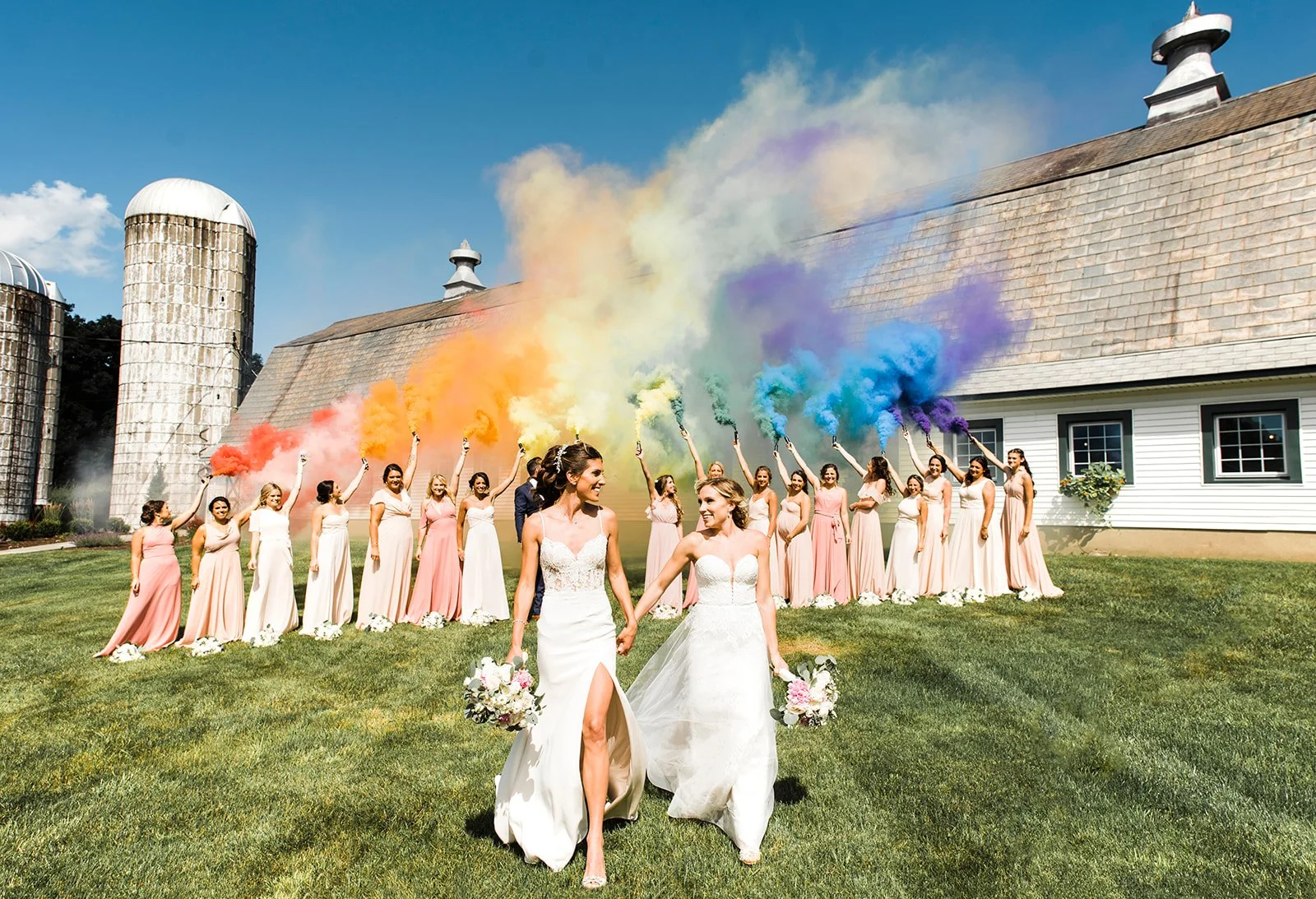 Two brides in wedding dresses holding hands walking on grass with a group of bridesmaids behind them holding colorful smoke grenades.