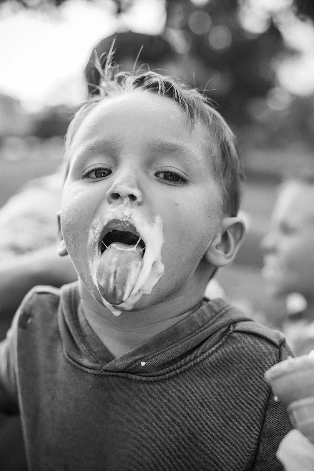 A young boy is licking ice cream with his mouth open, outdoors, with a blurred background.
