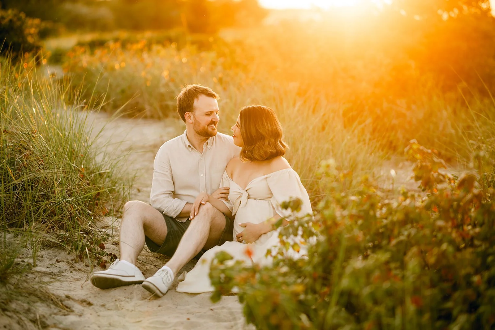 A couple sitting on a sandy path surrounded by tall grass and plants during sunset, looking at each other affectionately.