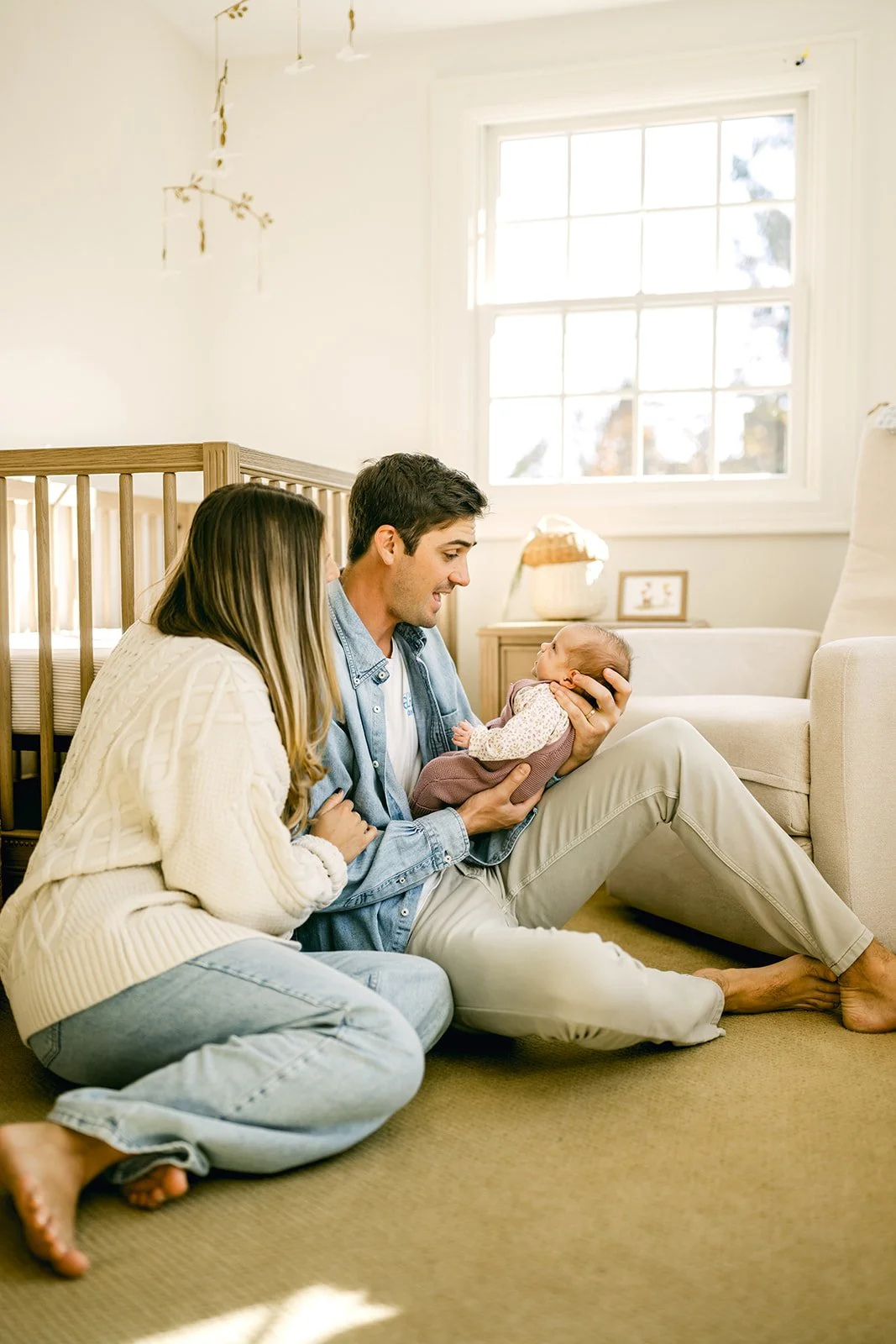 A family of three sitting on the floor of a bright room with a large window, a crib, and a nightstand with a lamp and picture frame. The father is holding a newborn baby.