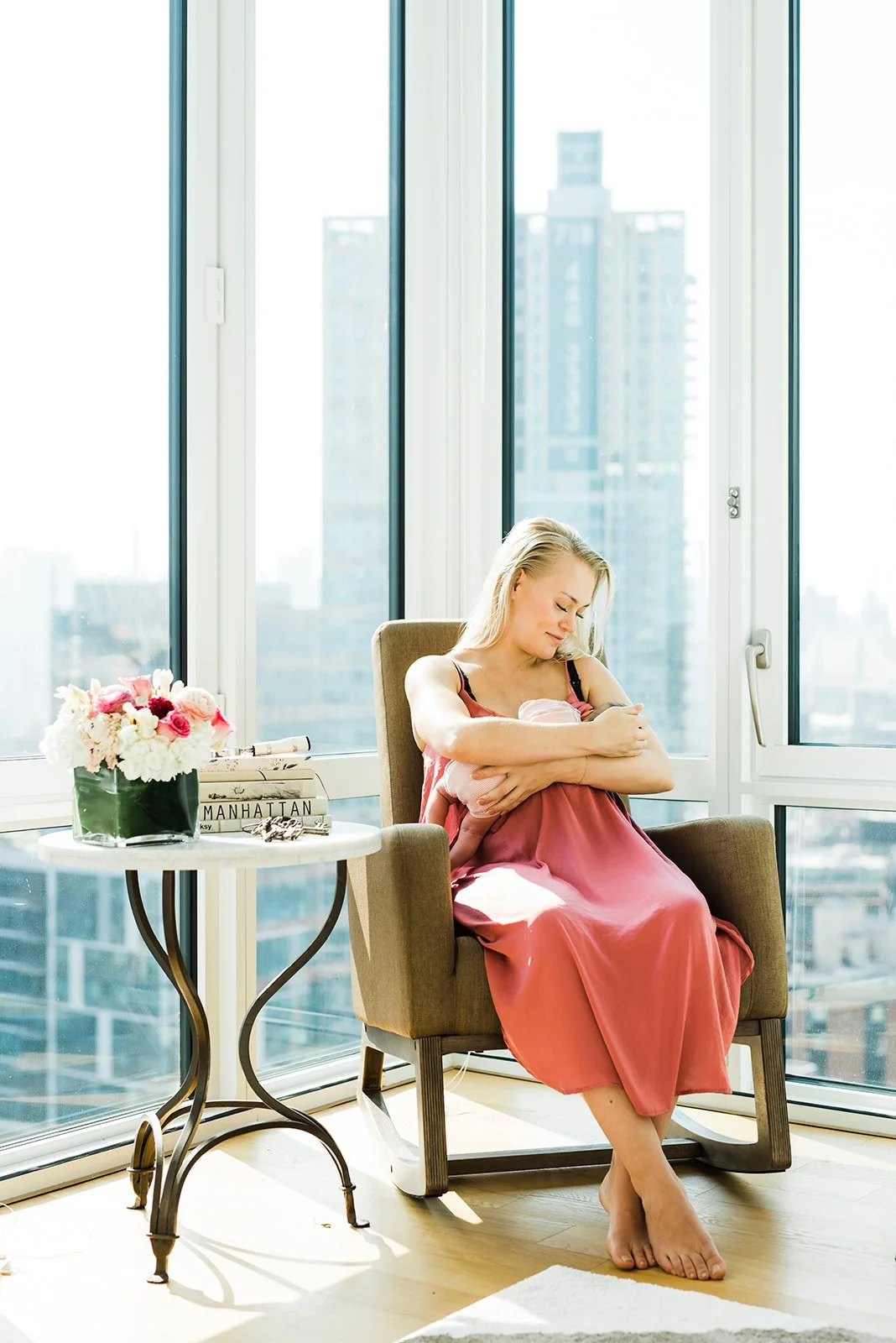 A woman in a pink dress breastfeeding a baby in a sunlit room with large windows showing city buildings outside
