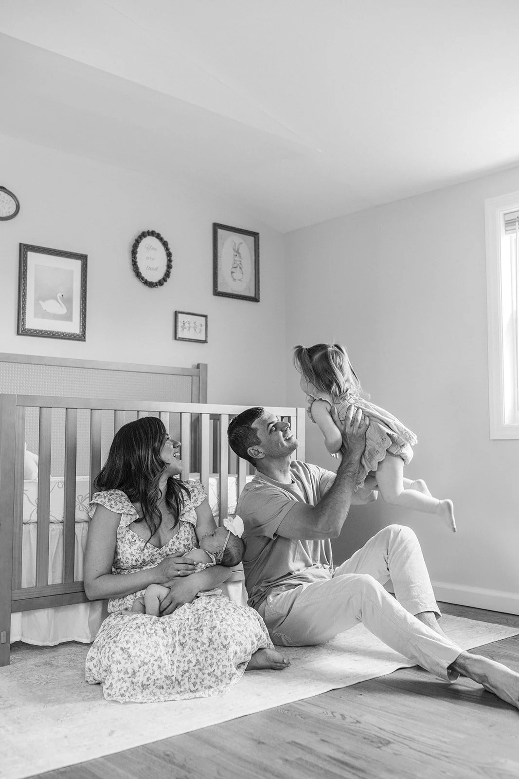 A family of three celebrating a moment in a nursery, with the father lifting a young girl while mother sits on the floor holding a baby, all smiling and enjoying the moment.