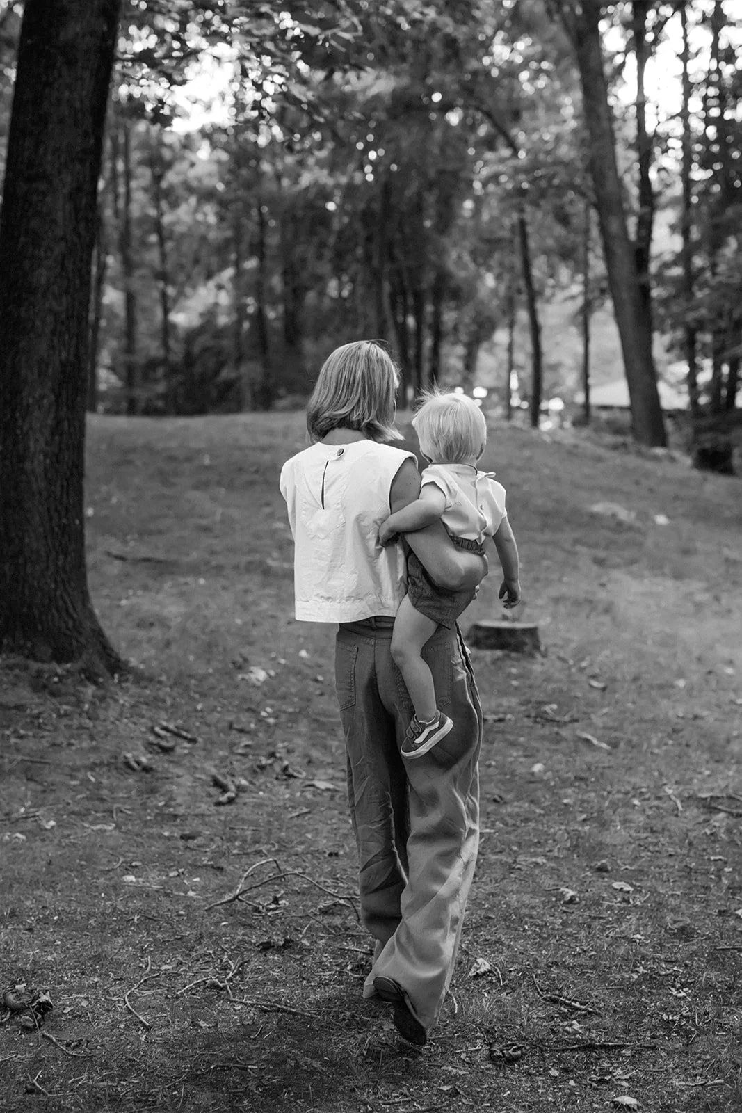 A woman walking outdoors through a wooded area with a young child in her arms.