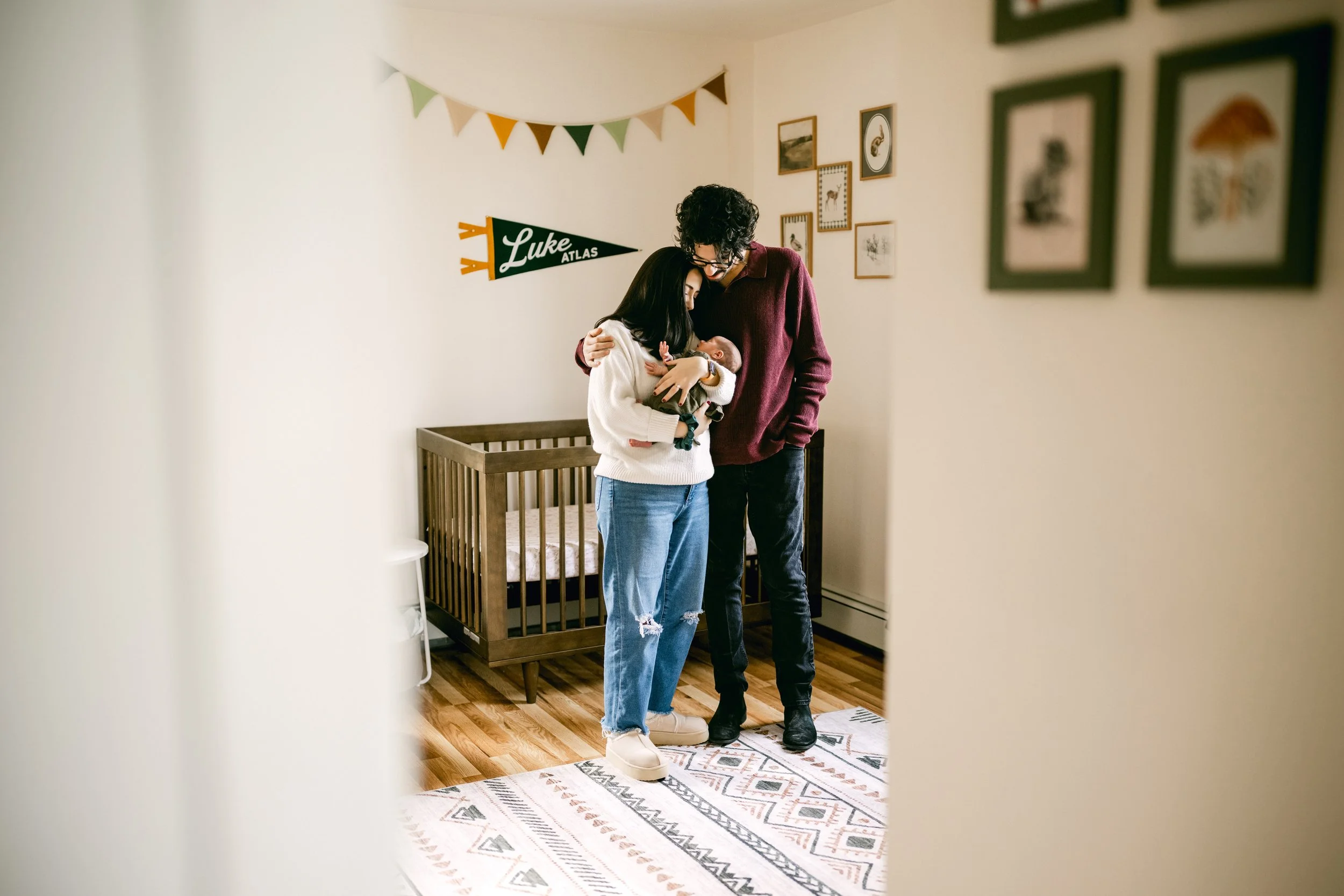 A couple, a woman and a man, are holding a newborn baby, standing in a nursery. The woman is wearing a white sweater and ripped jeans, the man a maroon sweater and black pants. The nursery has a wooden crib, framed pictures, and a string of colorful 