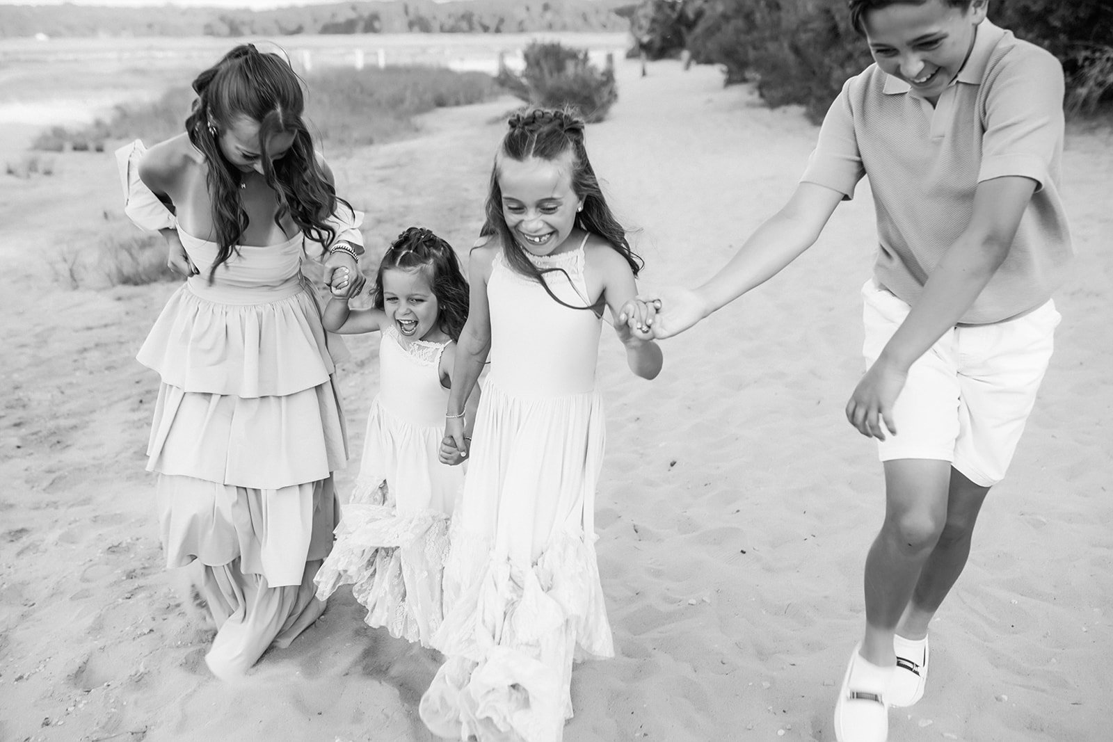 A group of five children, four girls and a boy, holding hands and playing on a sandy beach, all smiling and laughing.