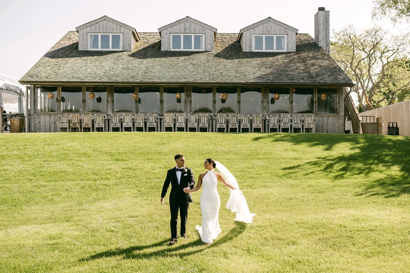 A bride and groom walking on a grassy lawn in front of a rustic wooden building with a porch full of chairs and hanging lanterns, on a sunny day.