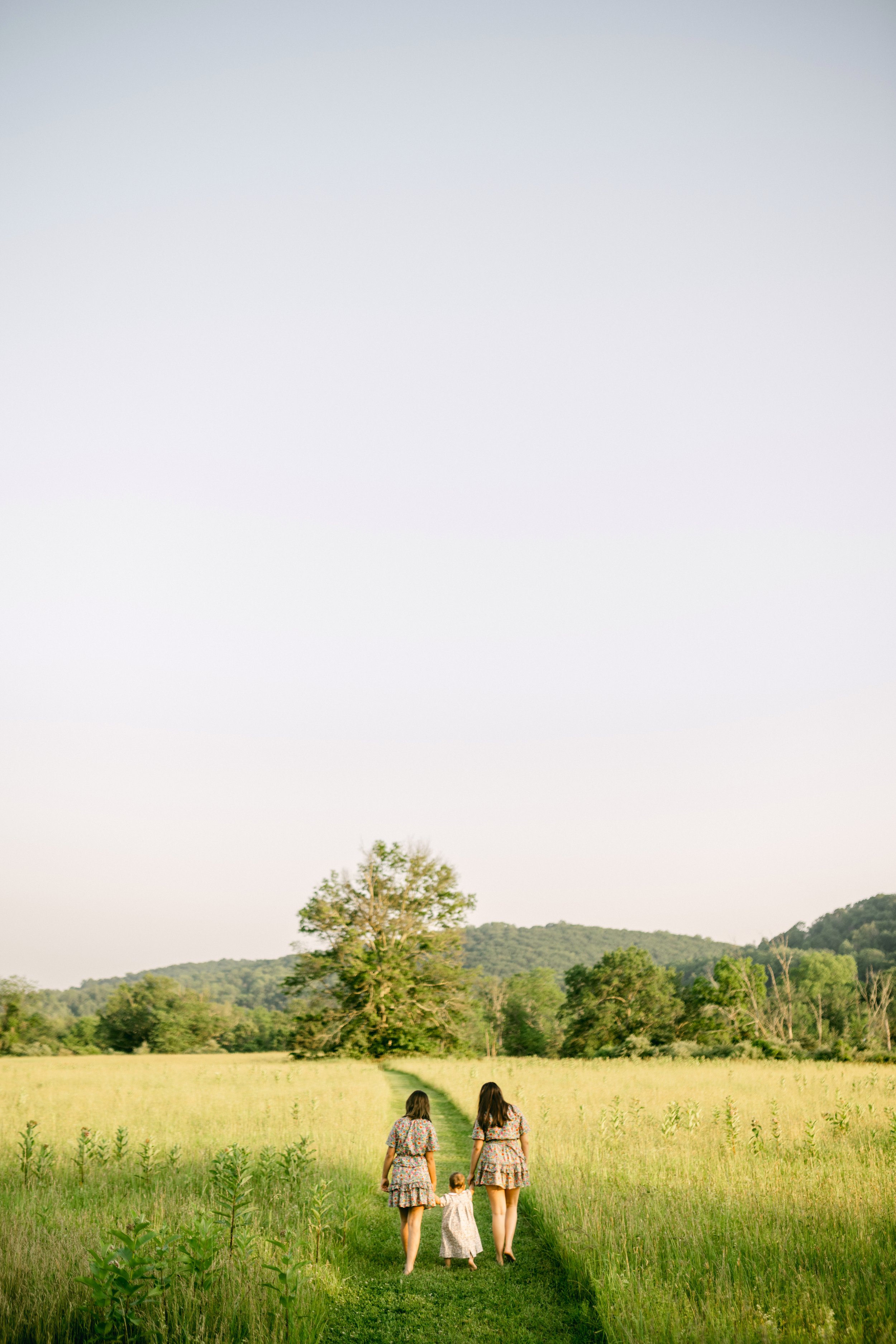 Two women and a young girl walk hand in hand down a grassy path through a green field, with trees and hills in the background under a clear sky.