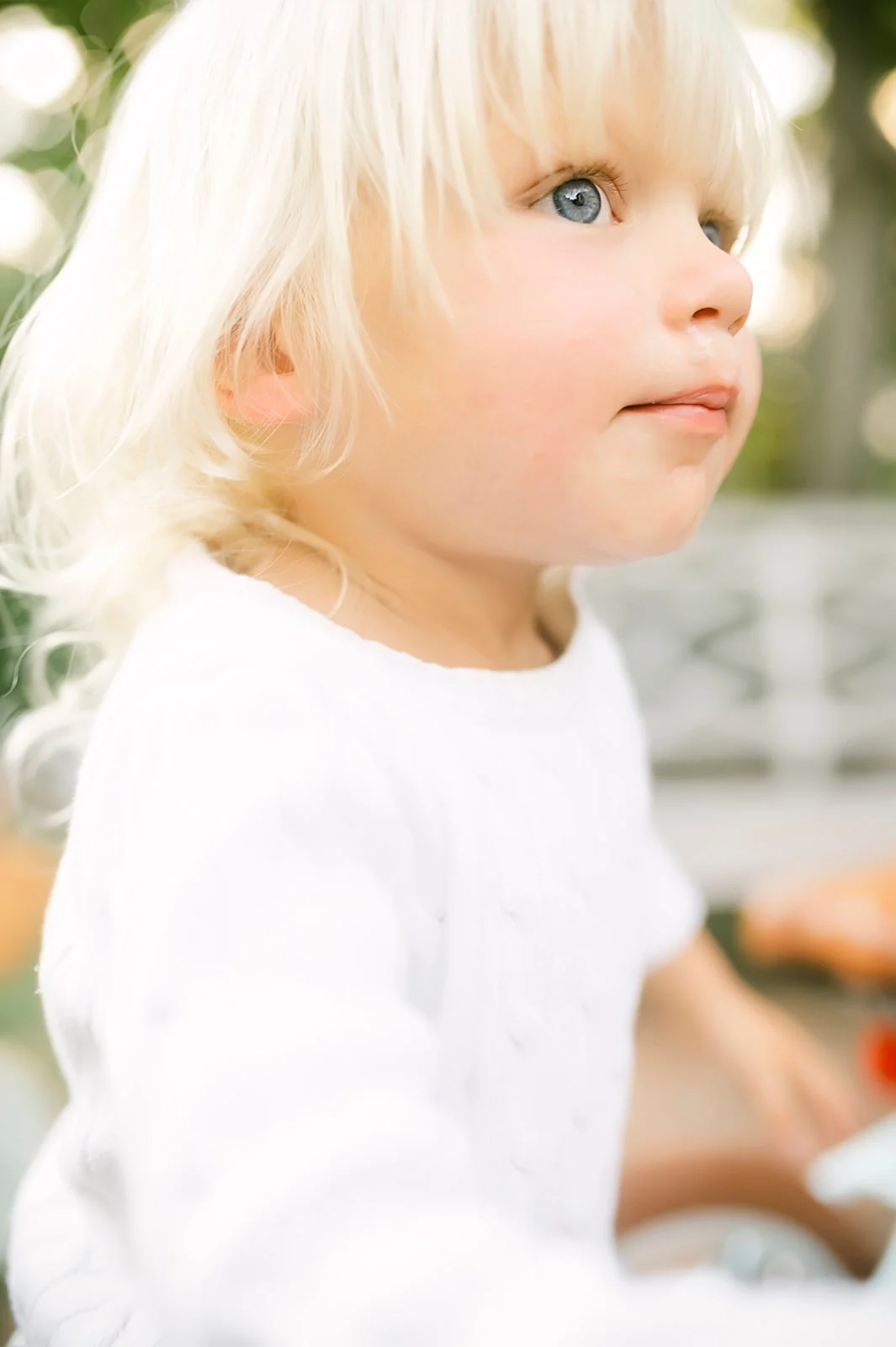 Close-up of a young blonde-haired girl with blue eyes wearing a white shirt, looking to the side outdoors.