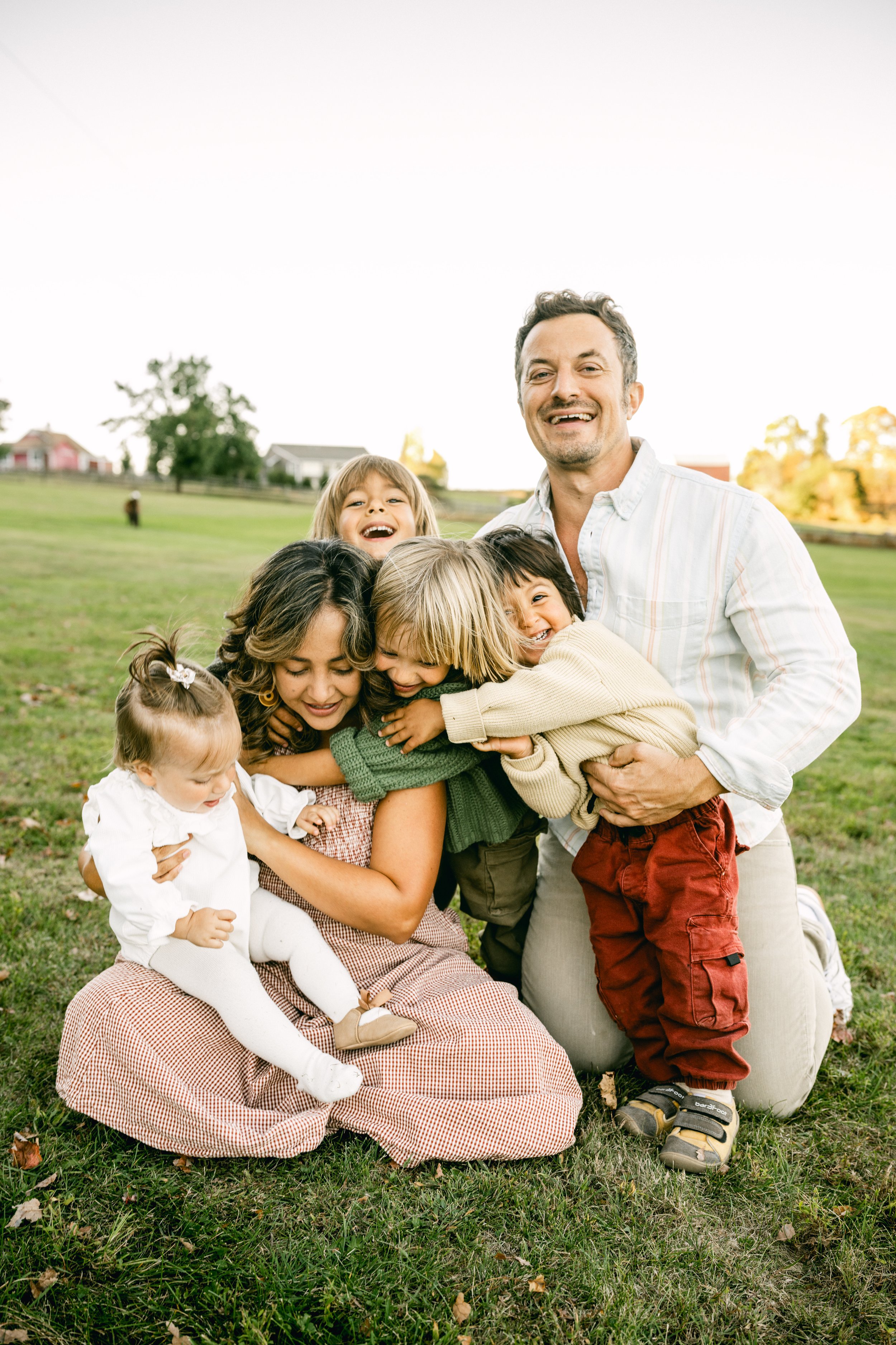 A family of six, including three children, hugging and smiling outdoors on a grassy field during daytime.