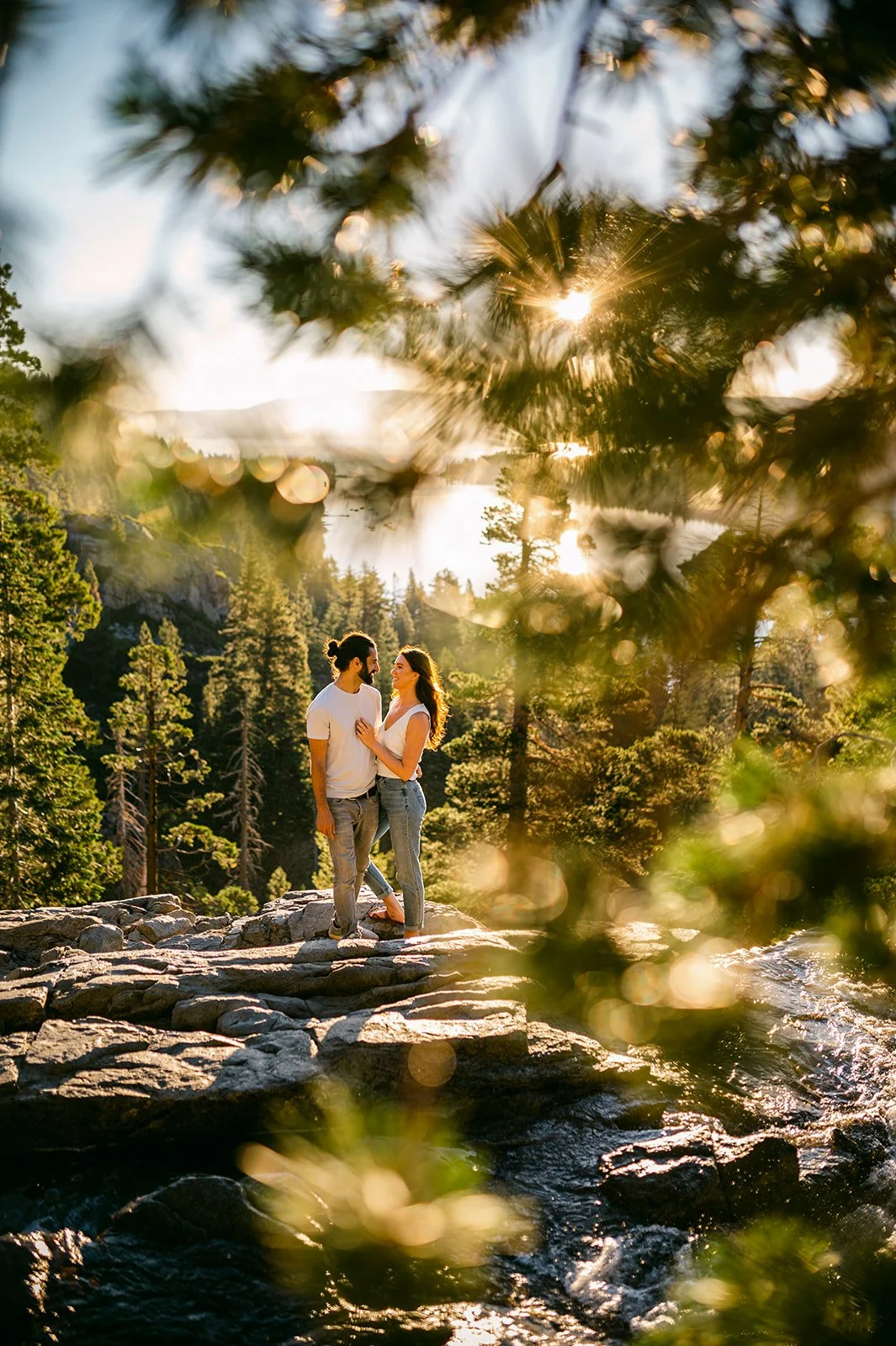 A couple stands on rocks near a river or waterfall in a forested area during sunset, with trees and mountains in the background and sunlight filtering through the branches.