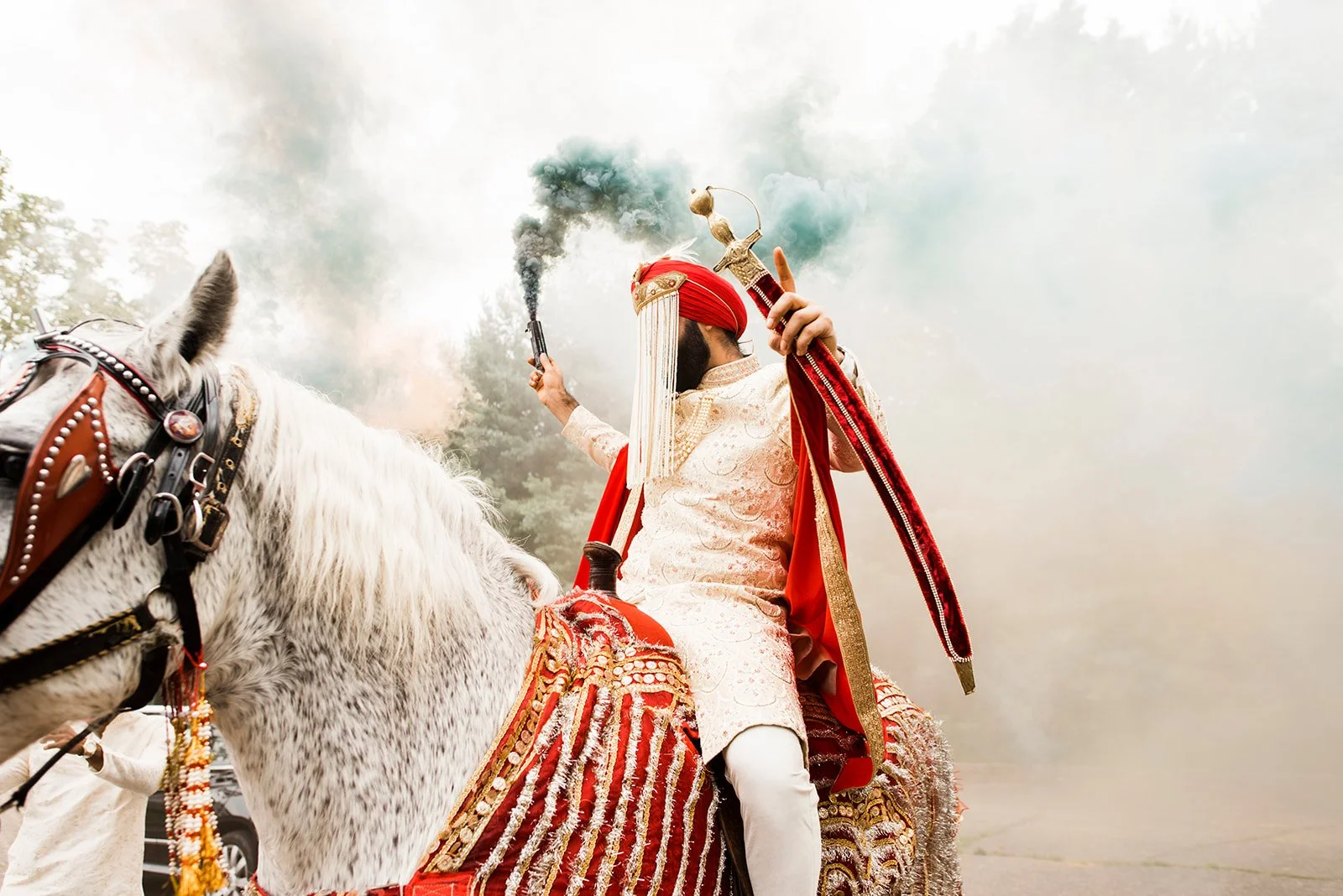 A man dressed in traditional Indian wedding attire riding a decorated white horse, holding smoke bombs and a sword, with a cloudy sky in the background.