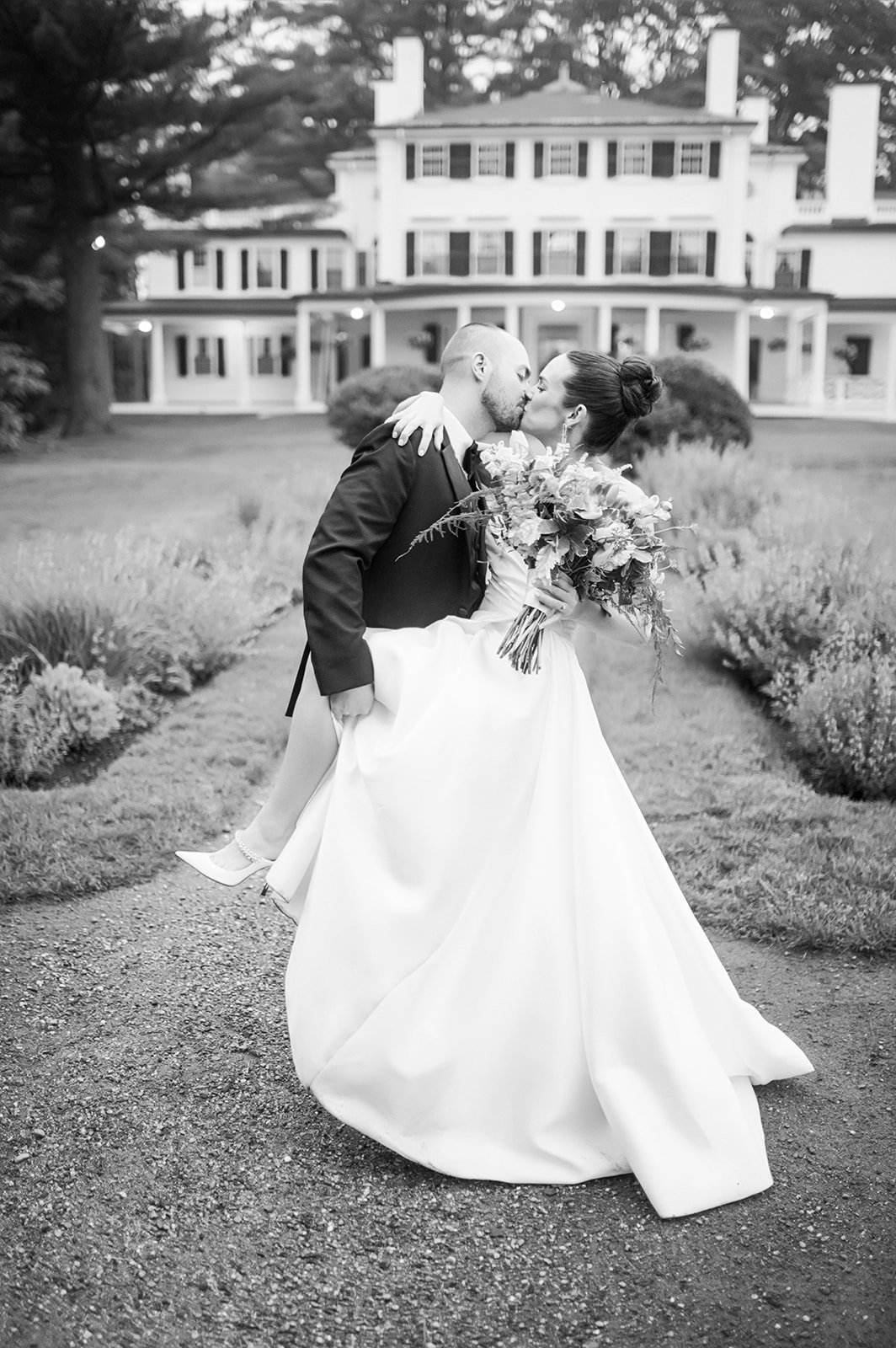 Black and white photo of a newlywed couple kissing. The groom carries the bride, holding a bouquet of flowers, in front of a large, historic house with a porch and multiple windows.