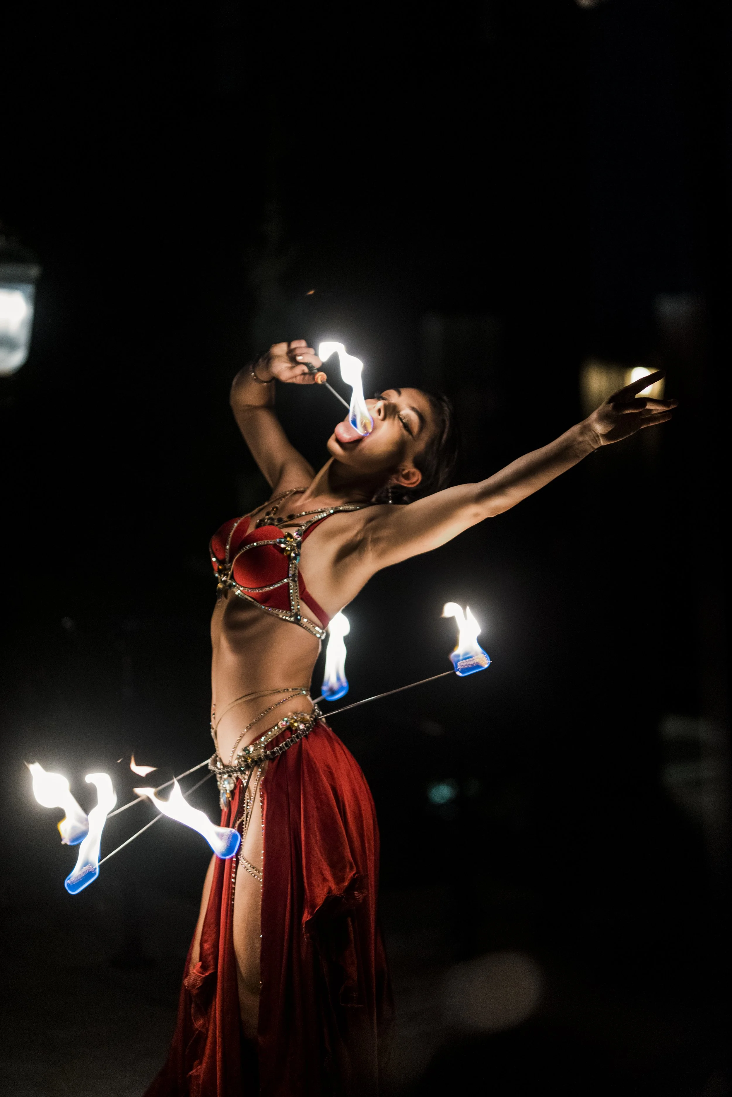 A fire performer dressed in a red costume with jewelry, twirling fire poi at night.