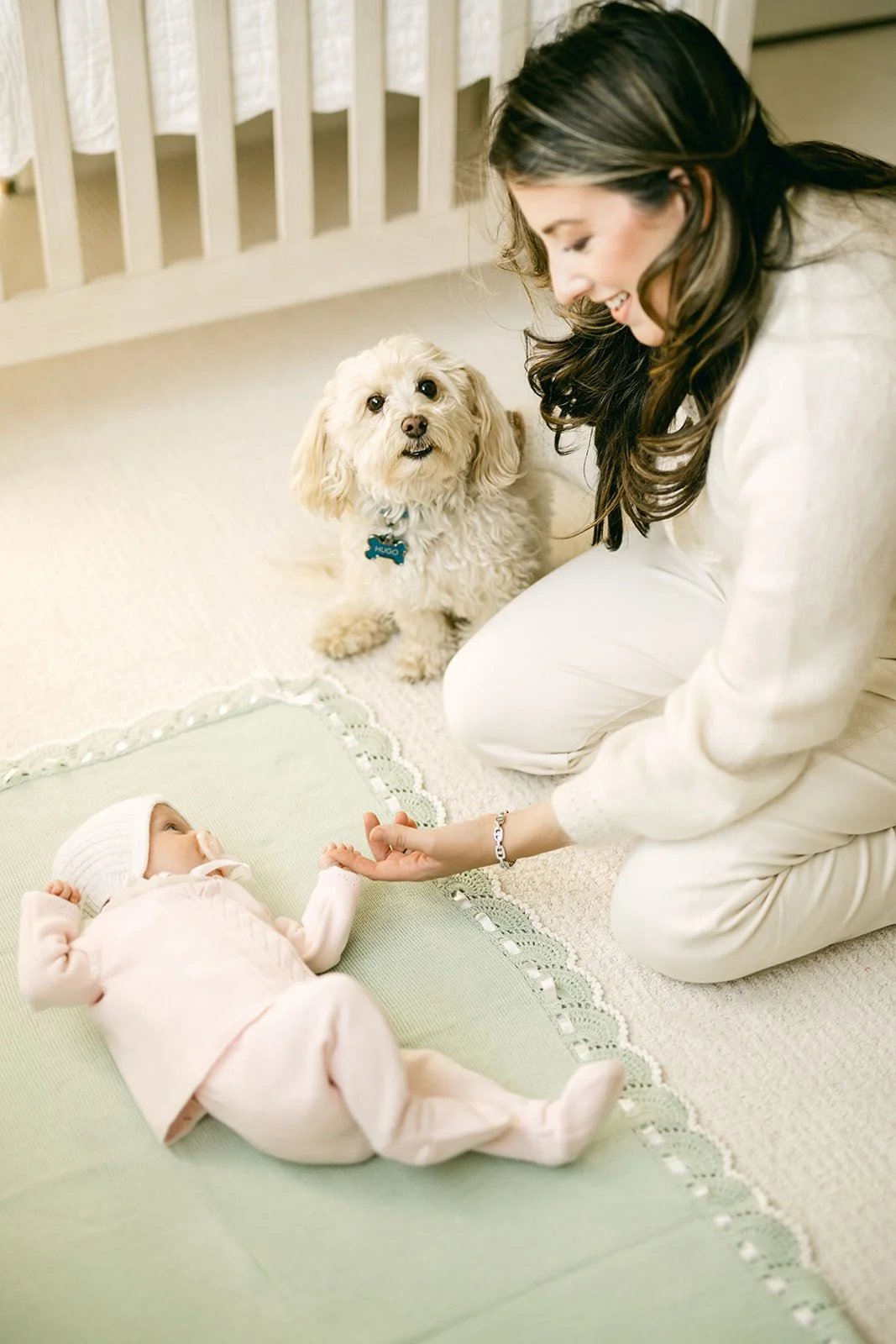 A woman crouching and holding a baby's hand, with a small fluffy dog nearby, inside a cozy room.