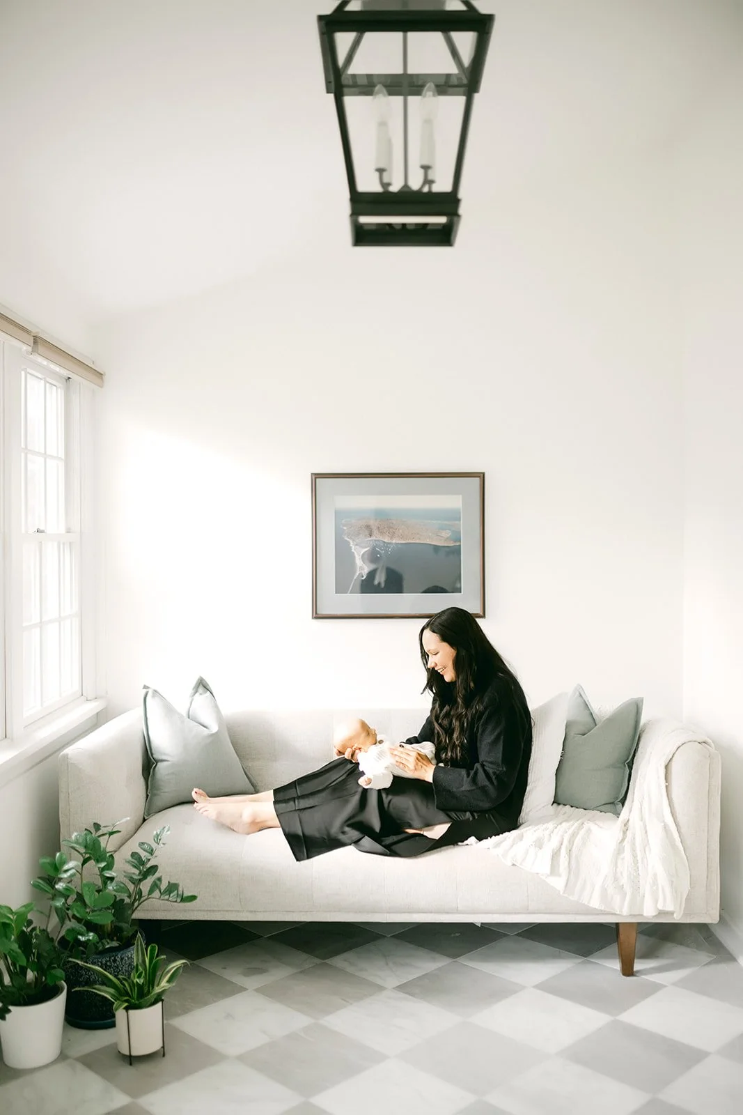 A woman in black pajamas sitting on a light-colored sofa holding a baby, smiling indoors near a window with a framed picture on the wall and potted plants nearby.