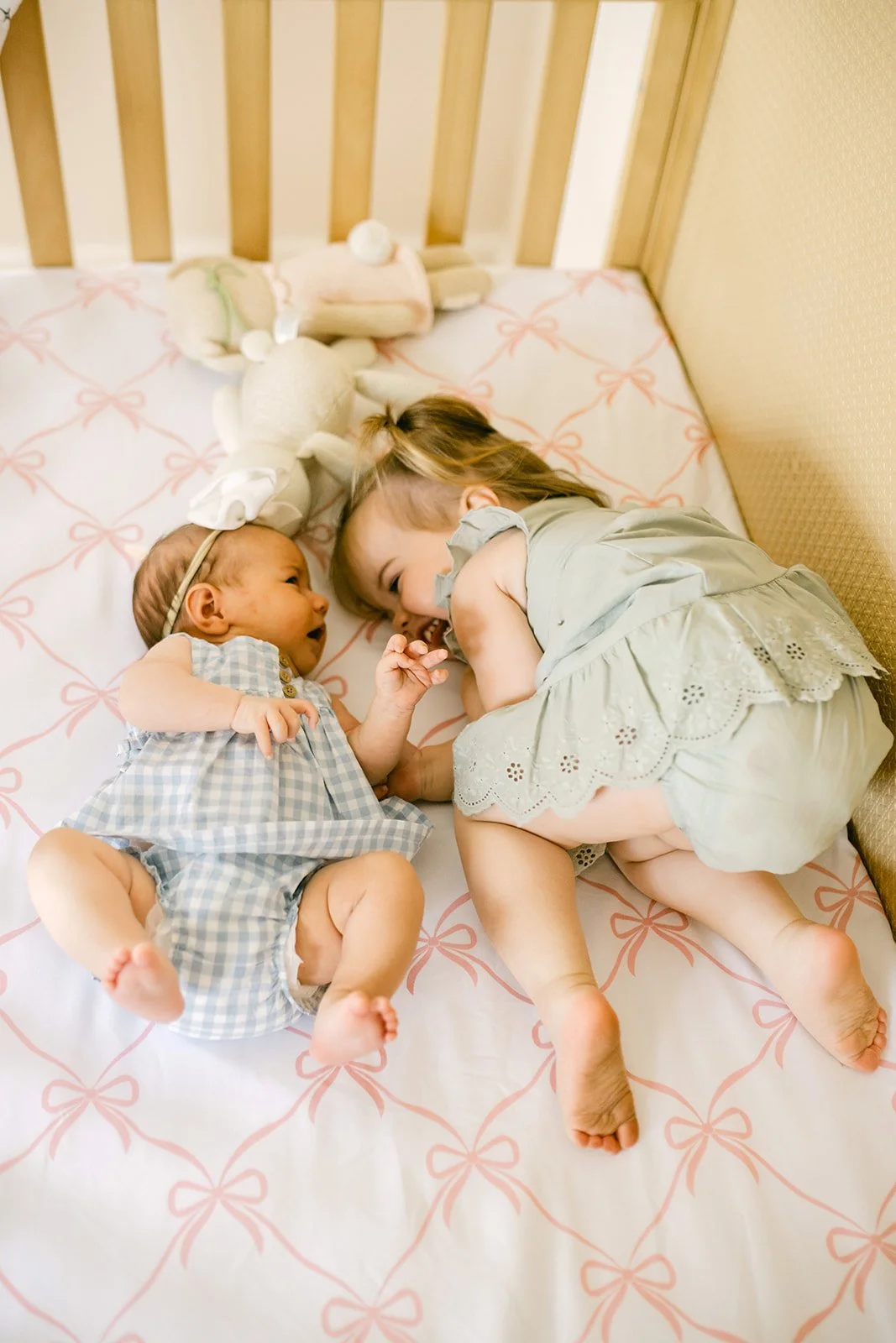 Two young girls, a toddler and a baby, are lying on a bed facing each other, smiling and touching hands. The bed has a white sheet with pink bow patterns, and there's a stuffed animal near them.