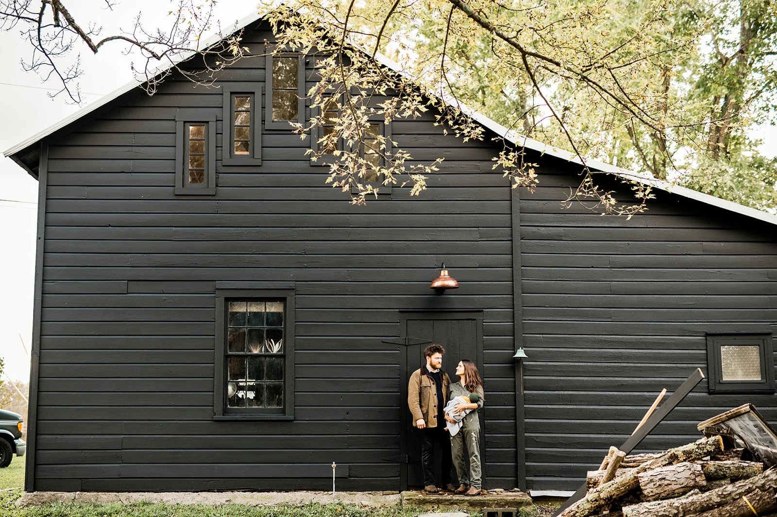 A black wooden barn with three small windows and a door, with two people, a man and a woman, standing outside holding a small dog, surrounded by trees with autumn leaves.