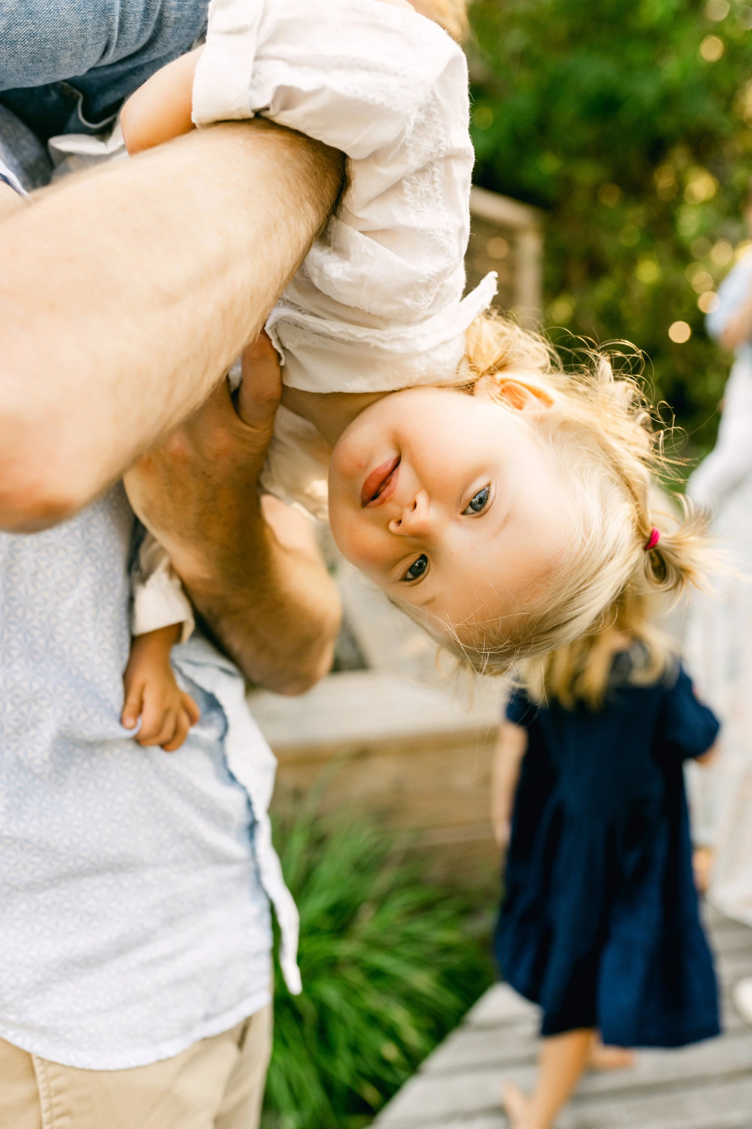 A man lifting a young girl upside down outdoors, with another child walking on a wooden path in the background.