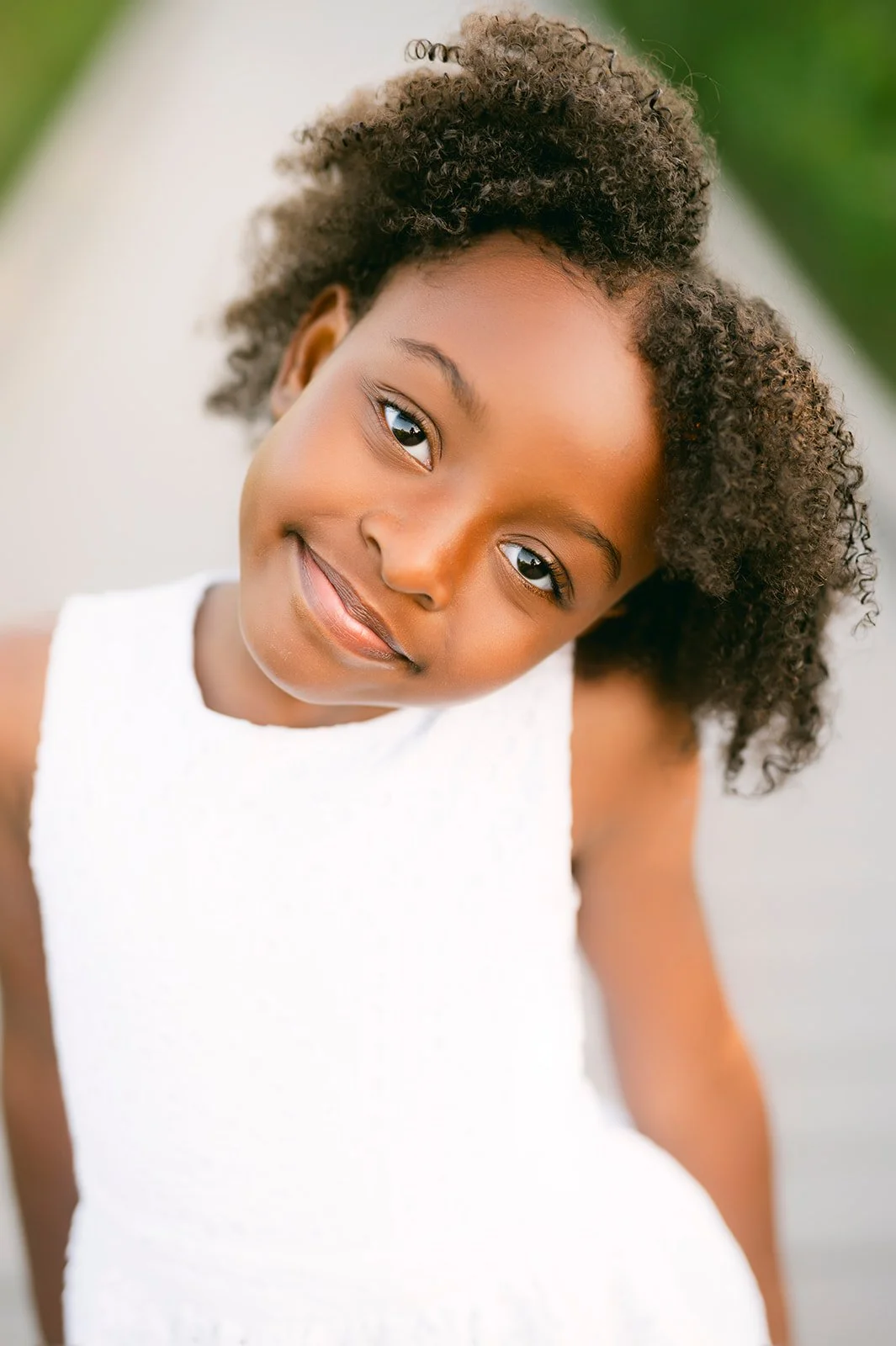 Close-up of a young girl with curly hair and brown skin wearing a white dress, smiling softly