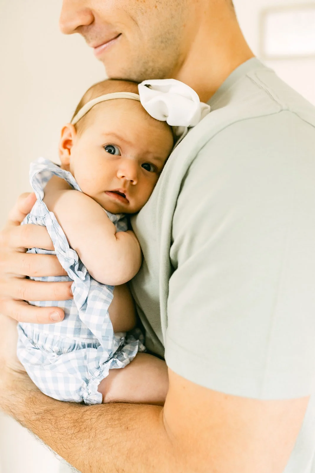 A man holding a baby girl with blue eyes, wearing a light blue gingham dress and a white bow headband, close to his chest, with both looking at the camera.