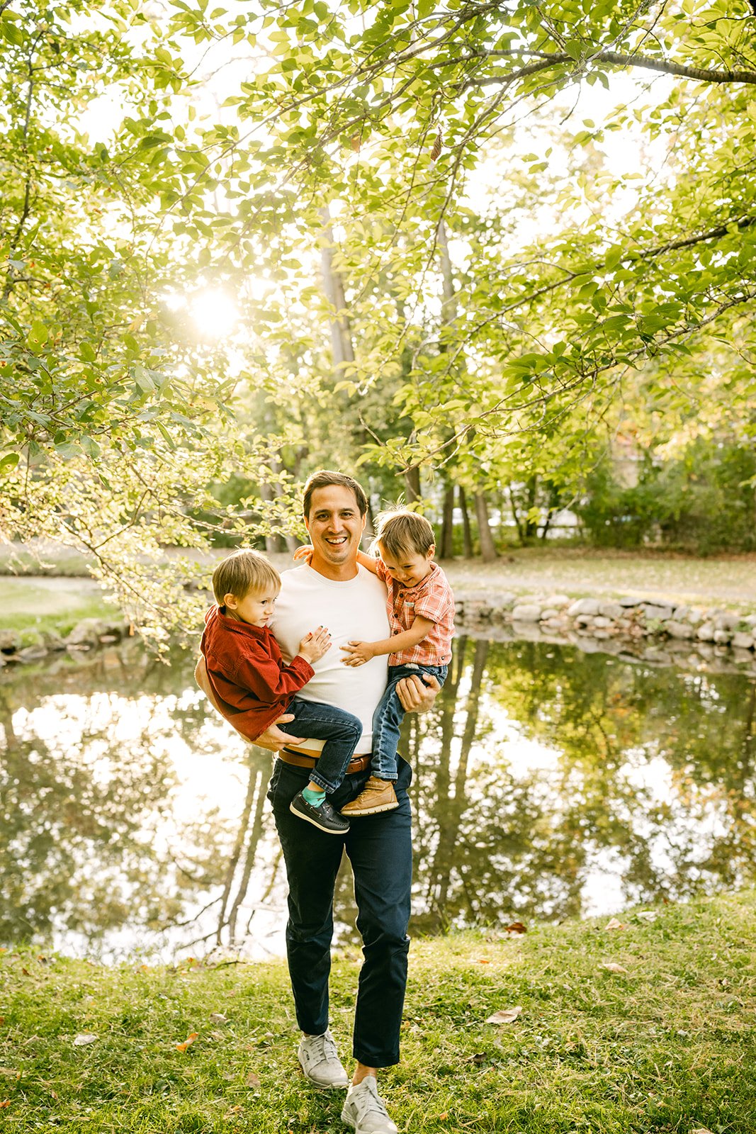 A man is walking outdoors on a grassy area near a pond, carrying two young children, one on each arm, with green trees and sunlight in the background.