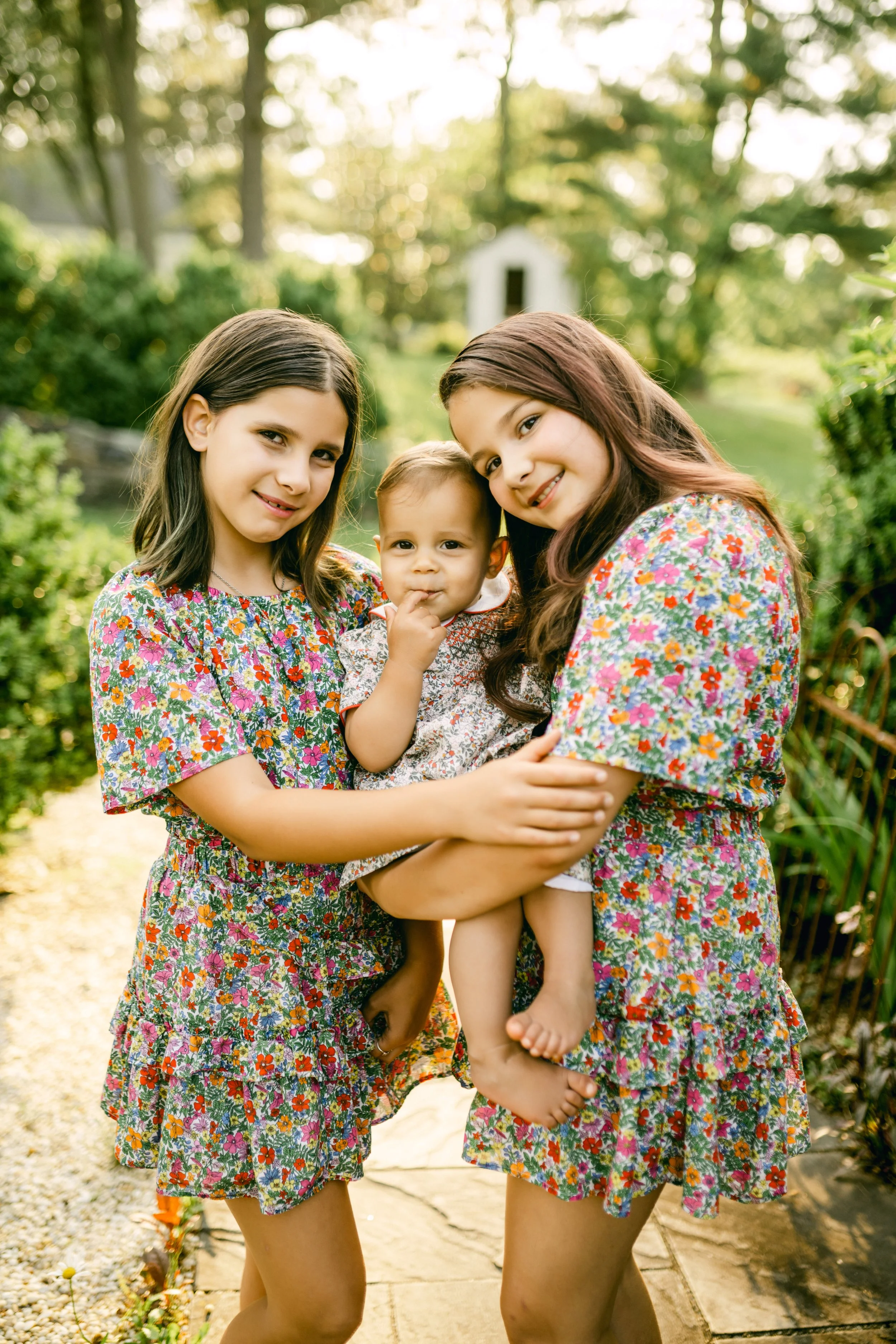Two women holding a baby girl outdoors in a garden, all wearing floral dresses, smiling at the camera.