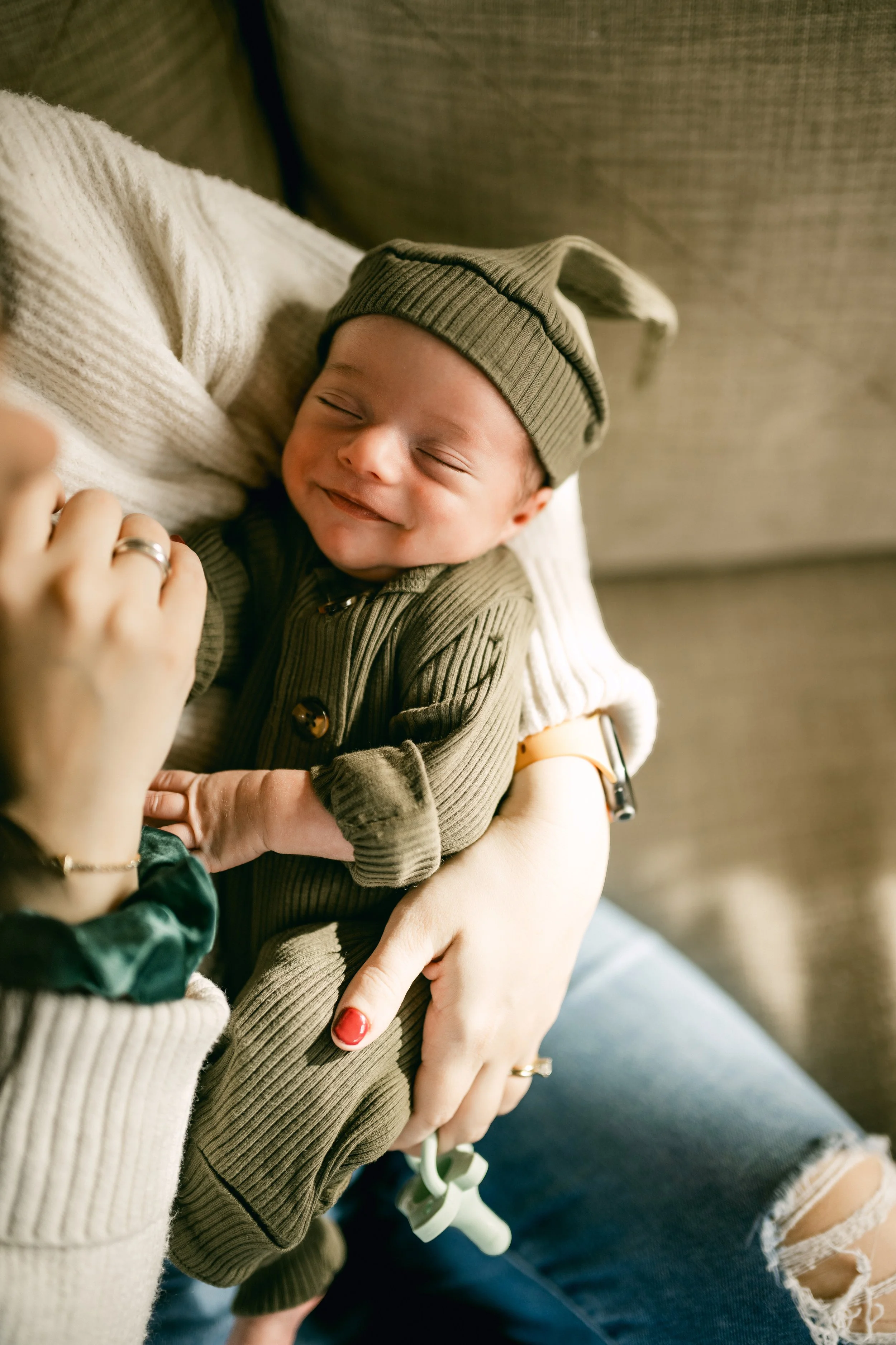 A smiling baby wearing a green hat and matching green outfit, being tenderly held by an adult with a beaded bracelet, sitting on a couch.