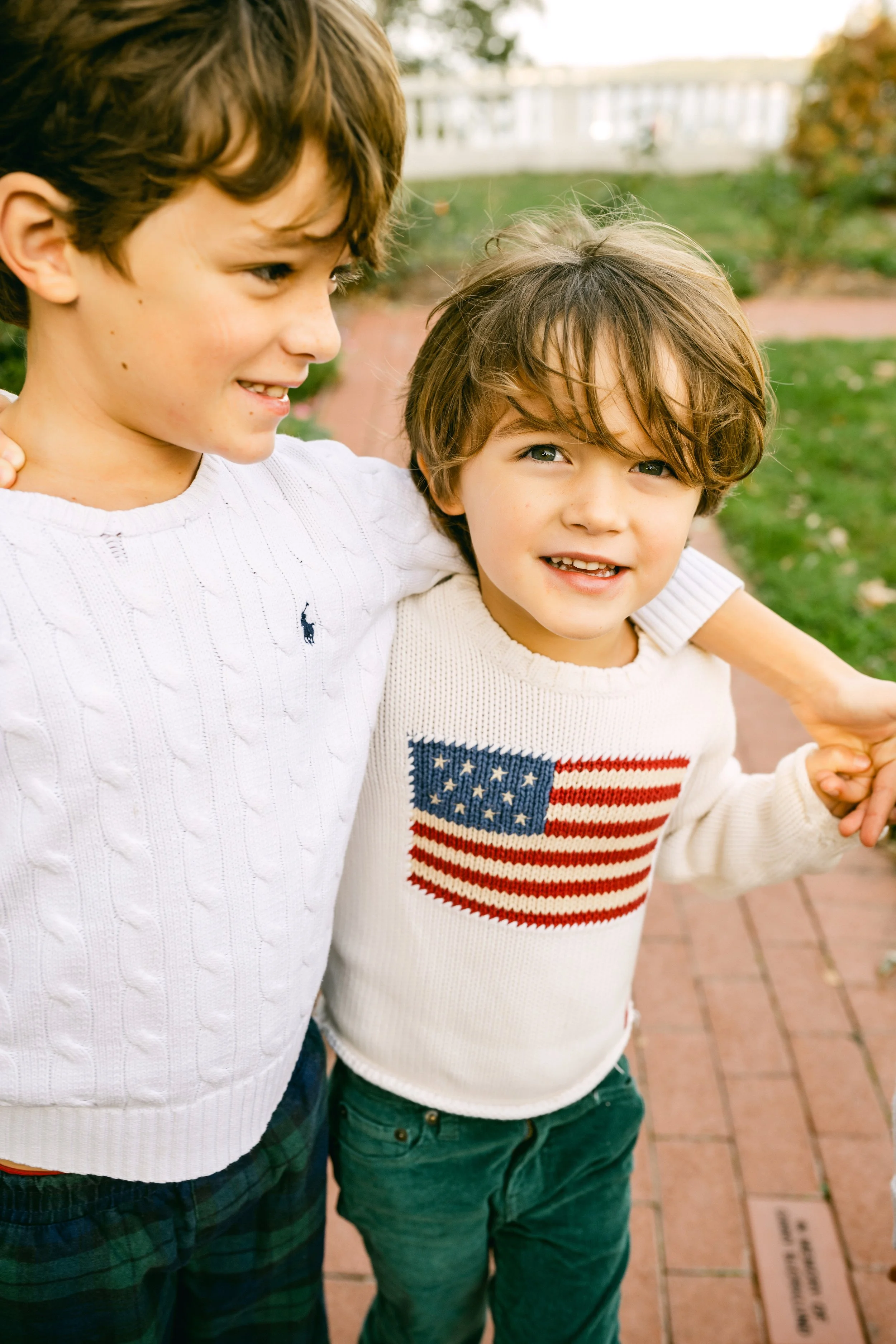 Two young boys standing outside with the older boy's arm around the younger boy, both smiling. The older boy is wearing a white sweater with a small dark logo, and the younger boy is wearing a white sweater with an American flag design. They are in a