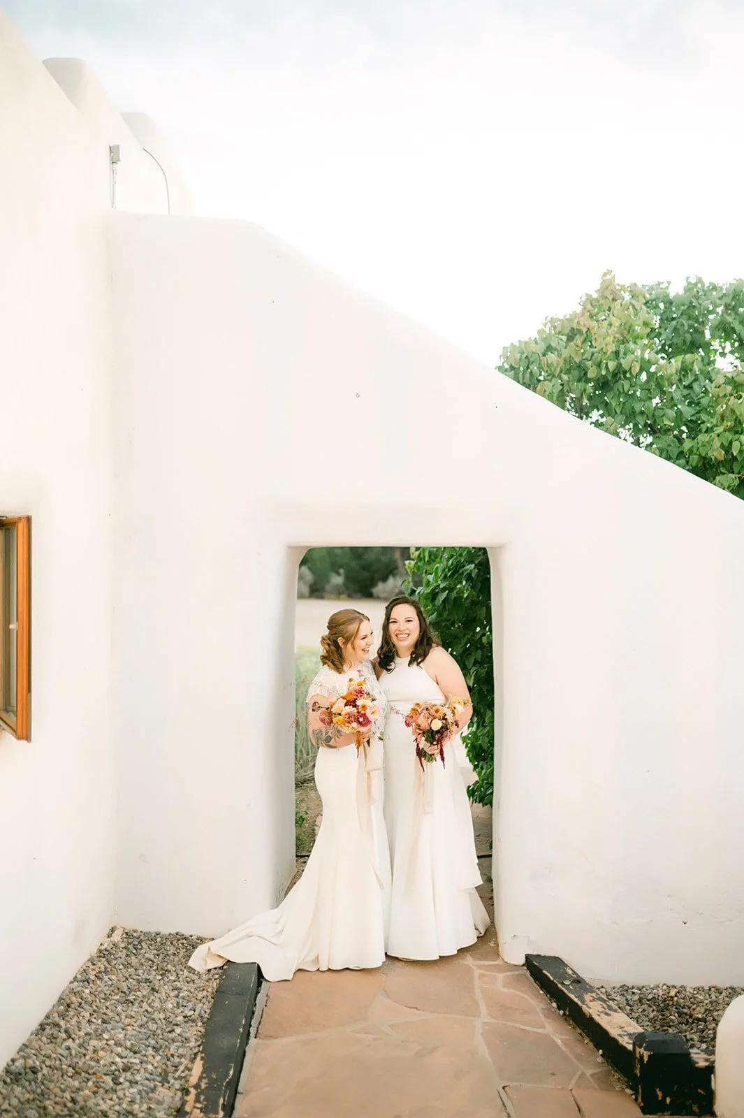 Two women in wedding dresses holding bouquets, standing close and smiling at each other in a white-walled outdoor setting.