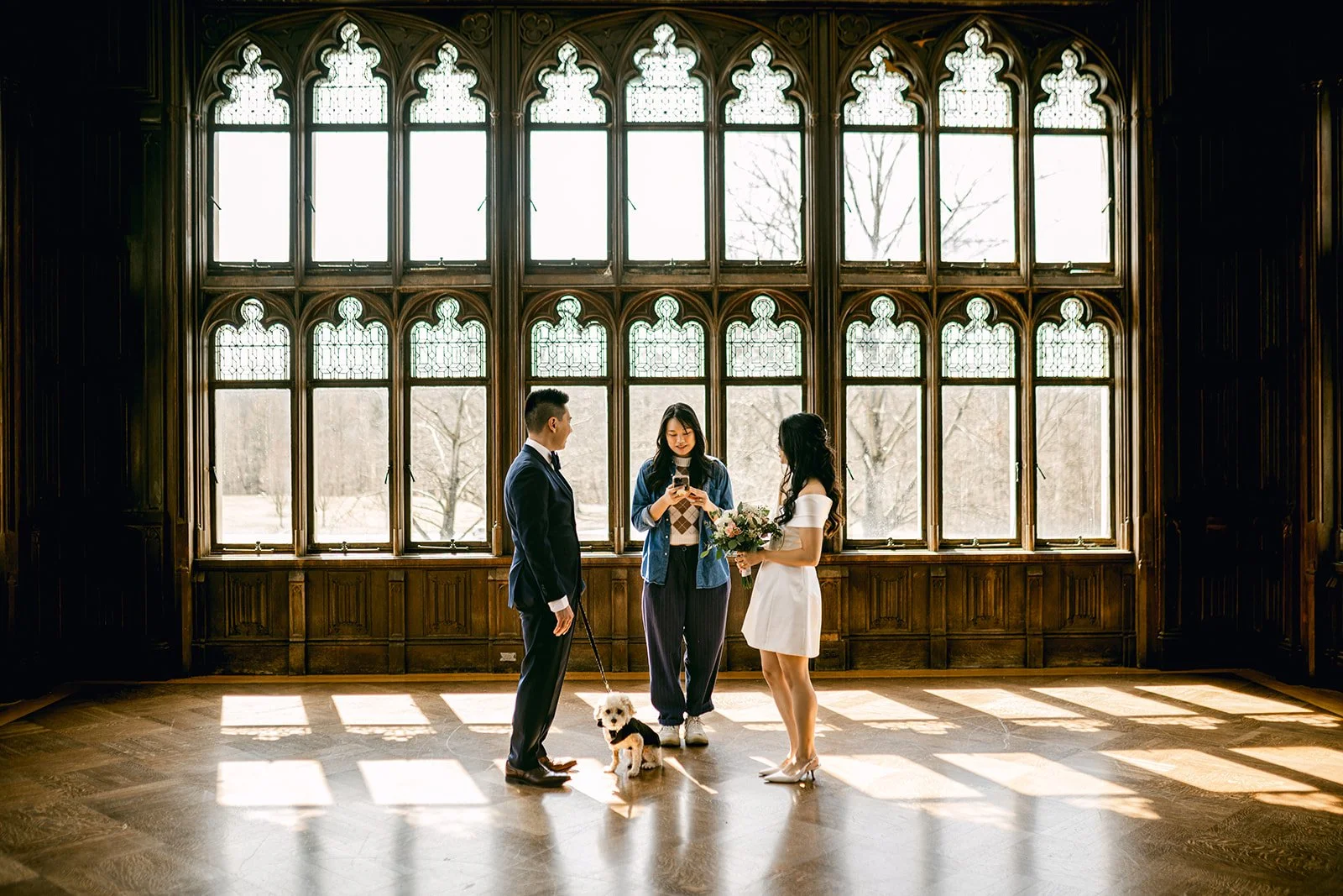 A wedding ceremony in a grand, wood-paneled room with large stained-glass windows. A bride in a white dress is holding a bouquet and standing with a groom in a suit, while a woman reads vows or a speech and another woman holding a dog dressed for the occasion looks on.