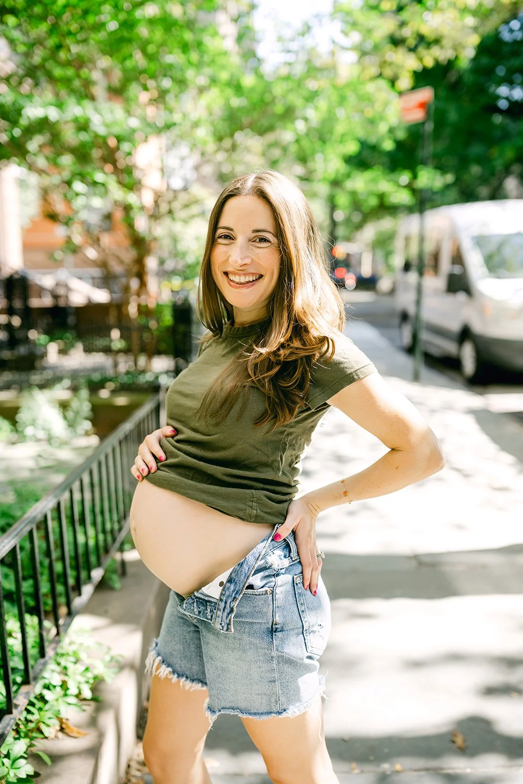 A pregnant woman with long brown hair smiling outdoors on a sunny day, wearing a green t-shirt and denim shorts, standing on a sidewalk near a black fence and green trees.