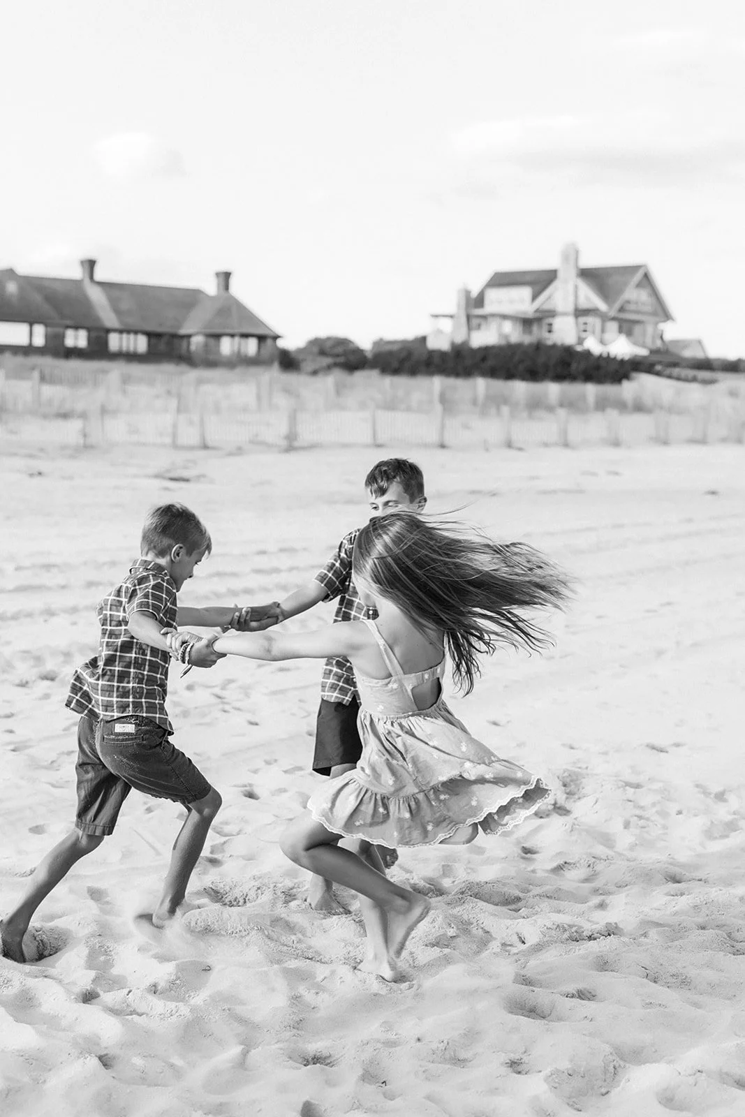 Three children playing and holding hands on a sandy beach, with houses in the background, black and white photograph.